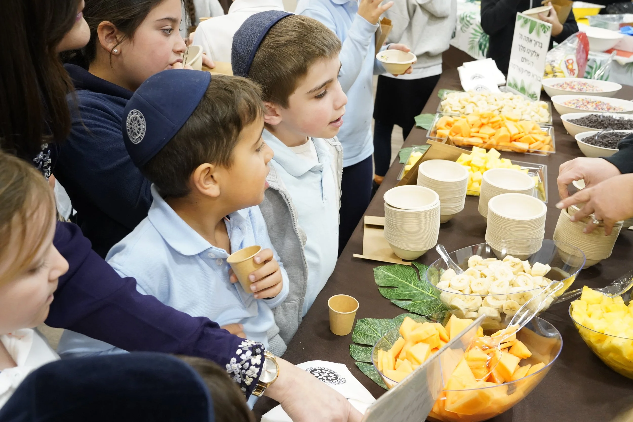 Children gathered around a table with bowls of sliced fruit and toppings, engaging in a food tasting event.