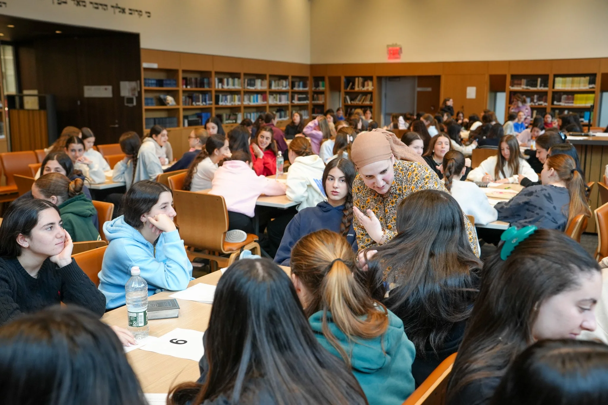 A large group of young women sitting at tables in a library or classroom setting, engaged in conversation. Some are looking towards a woman with a beige headscarf and a patterned shirt who is speaking to another woman. Shelves of books are visible in
