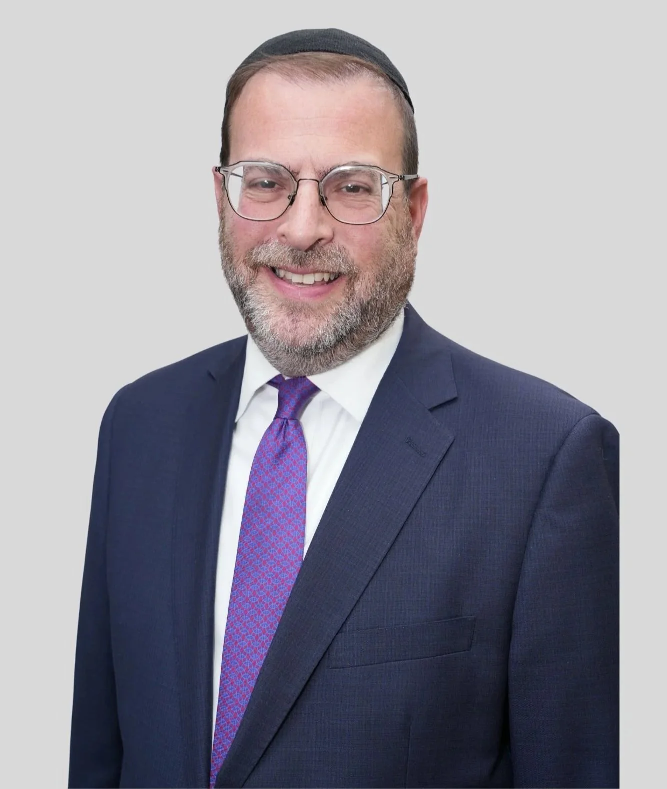 A middle-aged man with glasses, a beard, and a kippah, smiling and wearing a navy blue suit, white shirt, and patterned purple tie.