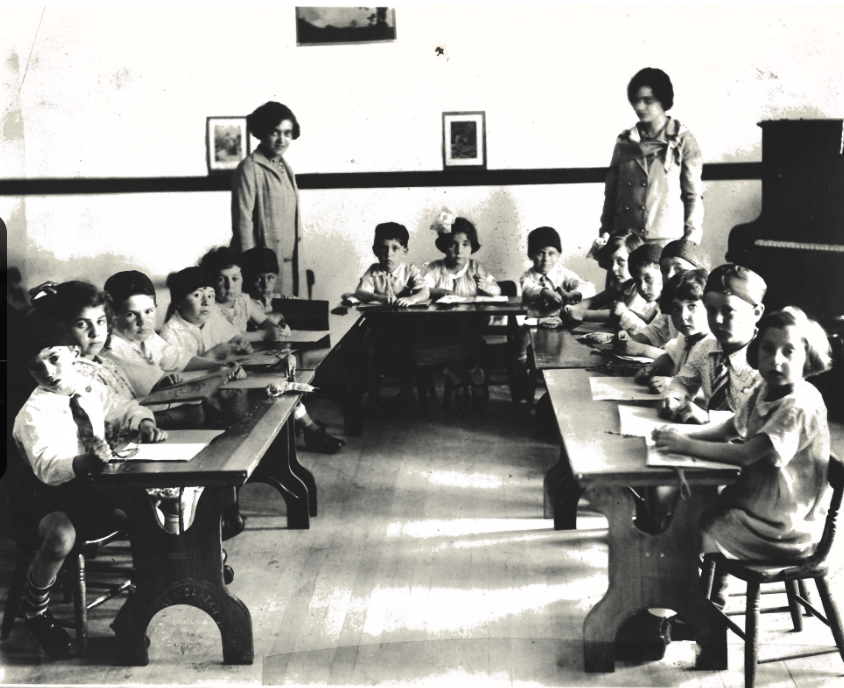 Black and white photo of a classroom with children sitting at desks, two women standing, and a piano in the corner.