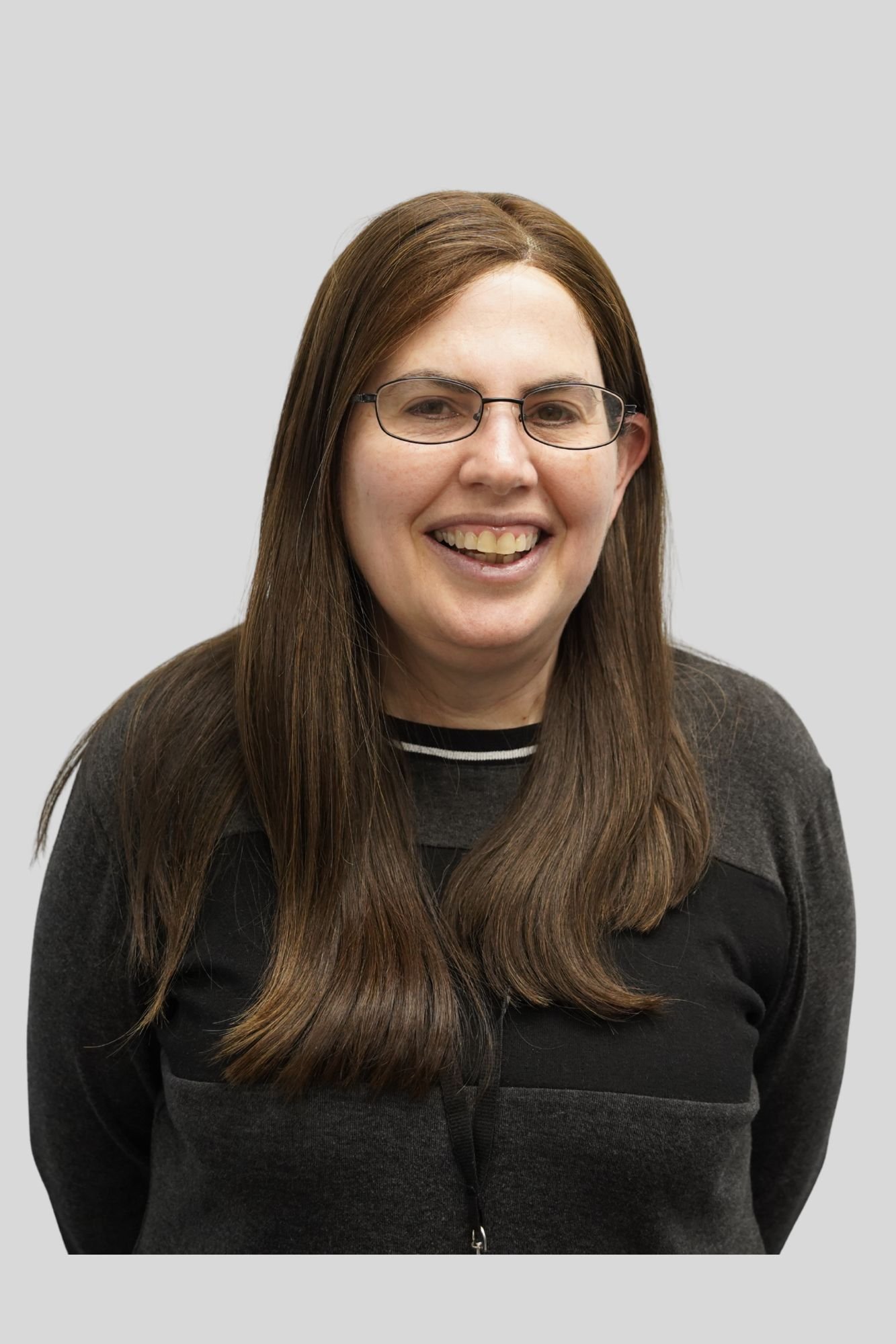 A woman with long brown hair, wearing glasses, a black and gray zip-up sweater, smiling with her teeth visible, standing against a plain light gray background.