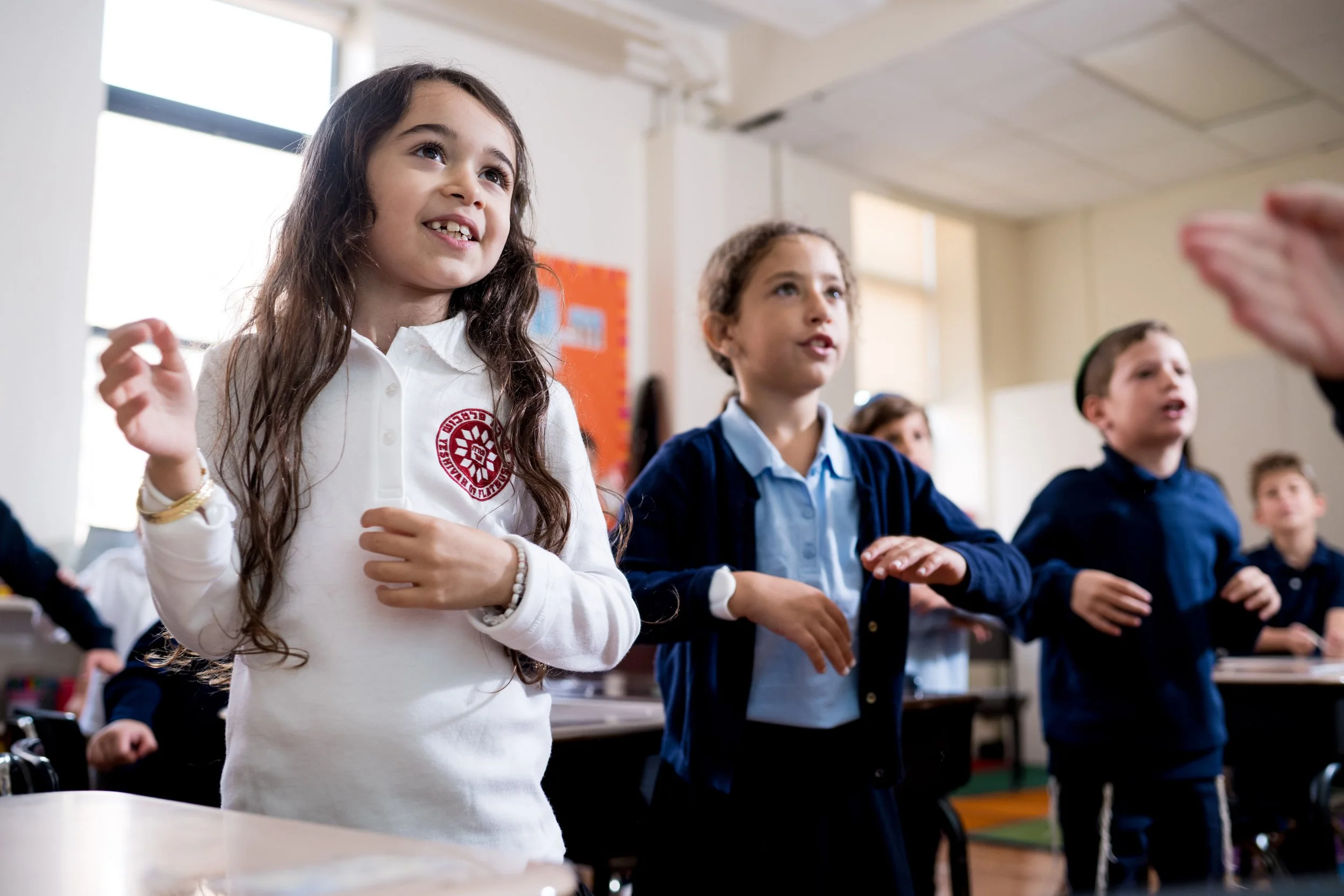 A classroom with young students standing and raising their hands, dressed in school uniforms, during an activity or lesson.