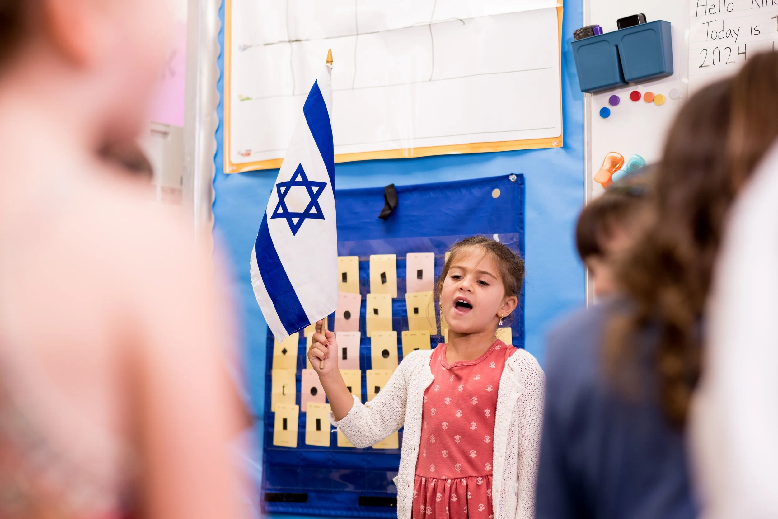 A young girl holding a small Israeli flag and speaking in a classroom with several children and colorful posters and a whiteboard in the background.