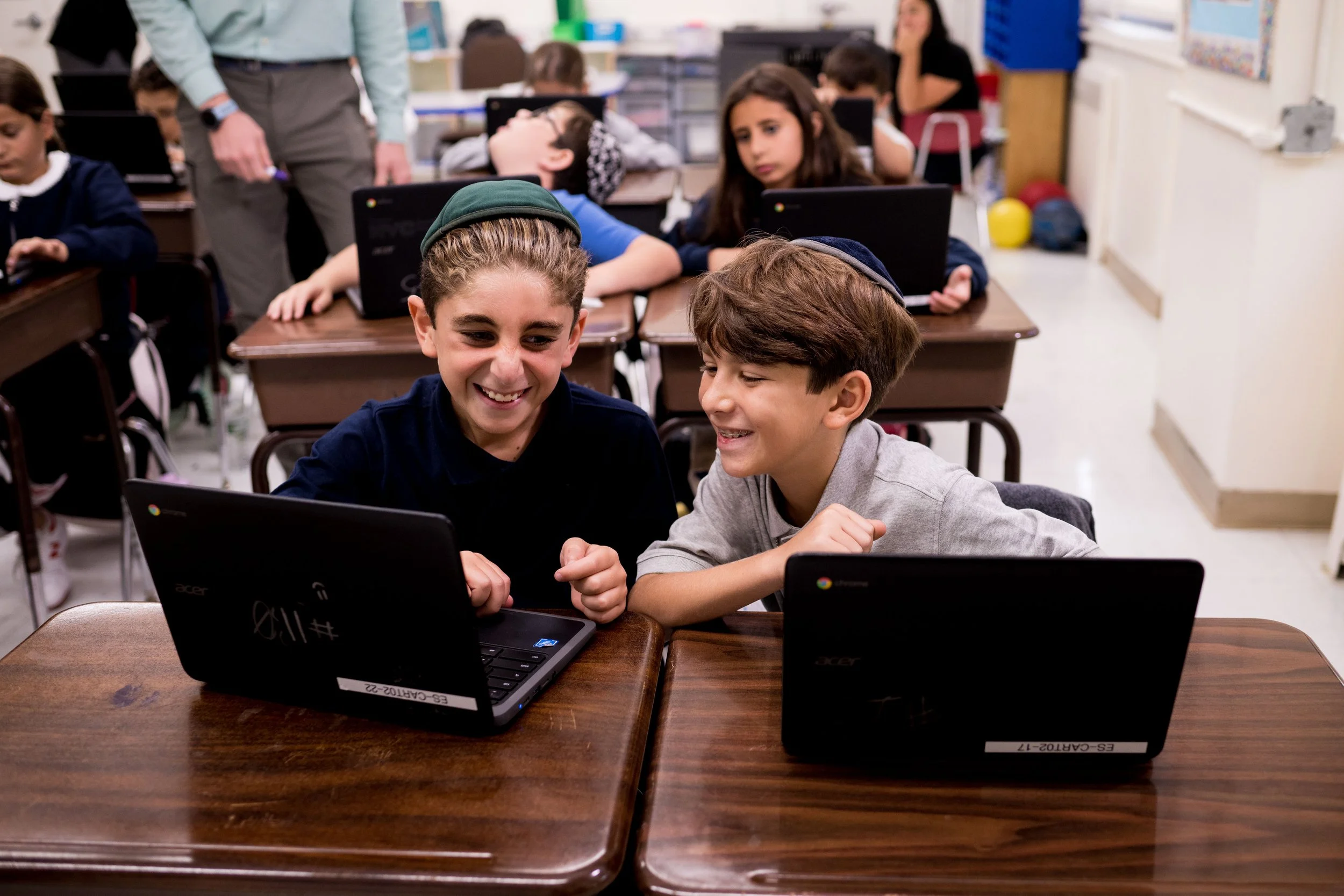 Two boys sitting at a wooden desk in a classroom, smiling and looking at a laptop, with other students and a teacher in the background.