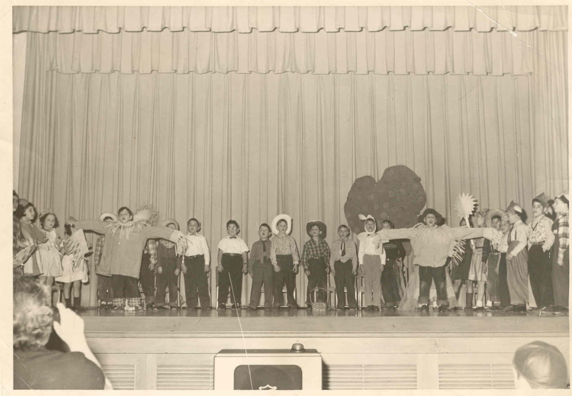 Children performing a school play with costumes and props on stage in front of an audience, with a curtain background.