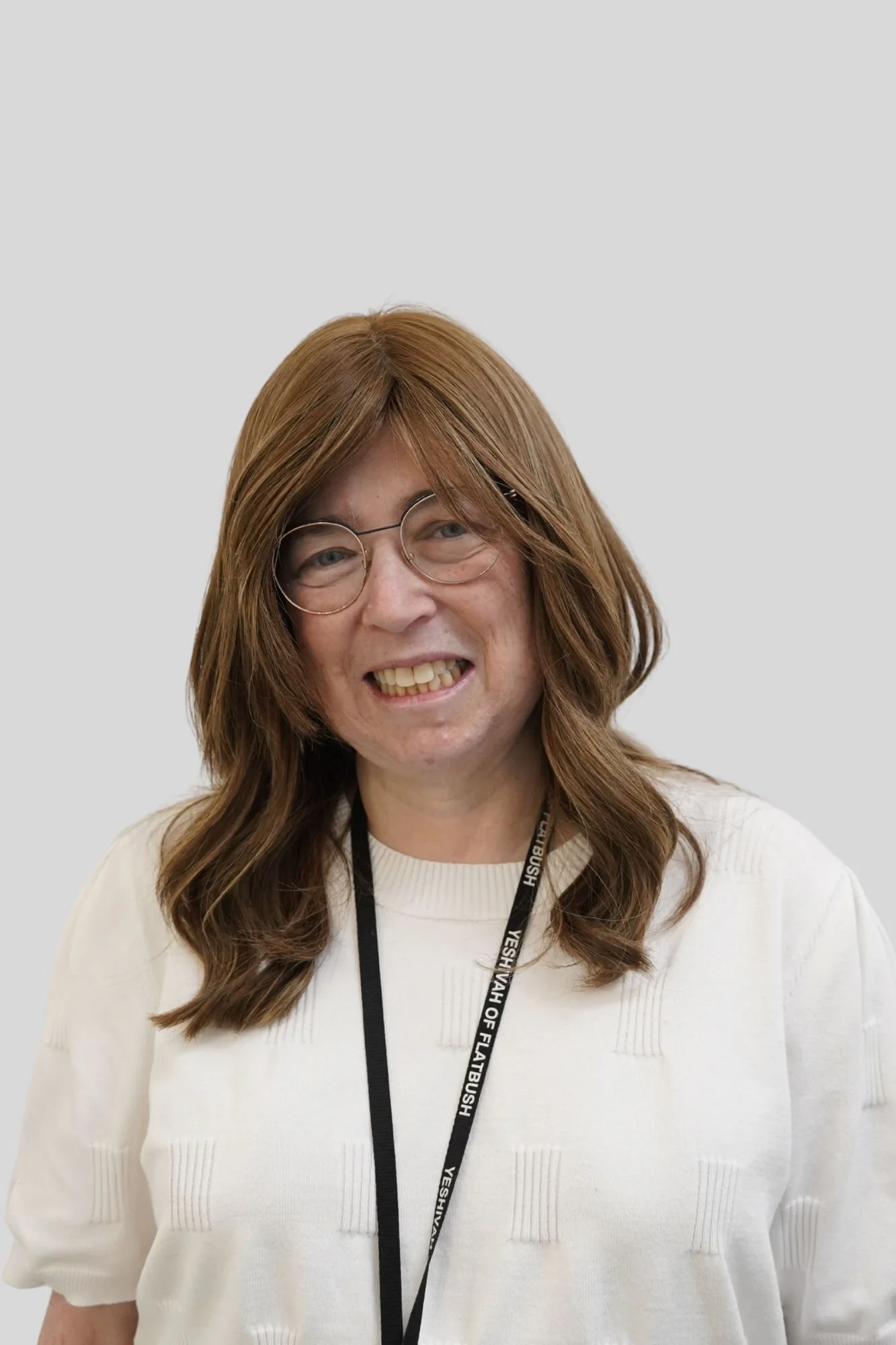 A woman with long, wavy, reddish-brown hair, wearing glasses and a white sweater. She has a name badge on a lanyard around her neck that reads "YESHIVA VAH OF FLATBUSH." She is smiling.