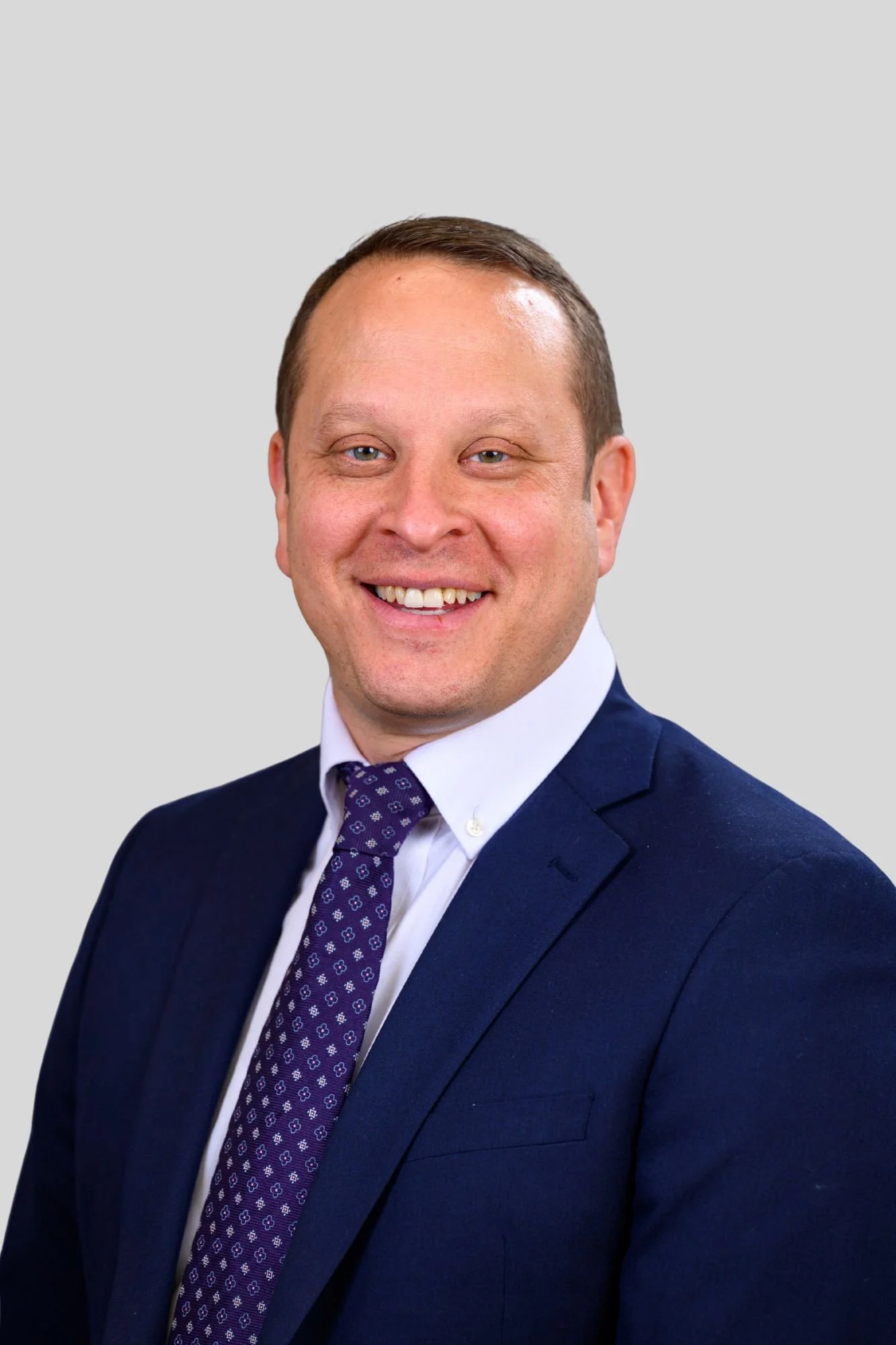 Portrait of a smiling man in a navy blue suit, white shirt, and purple tie with a pattern, against a light gray background.
