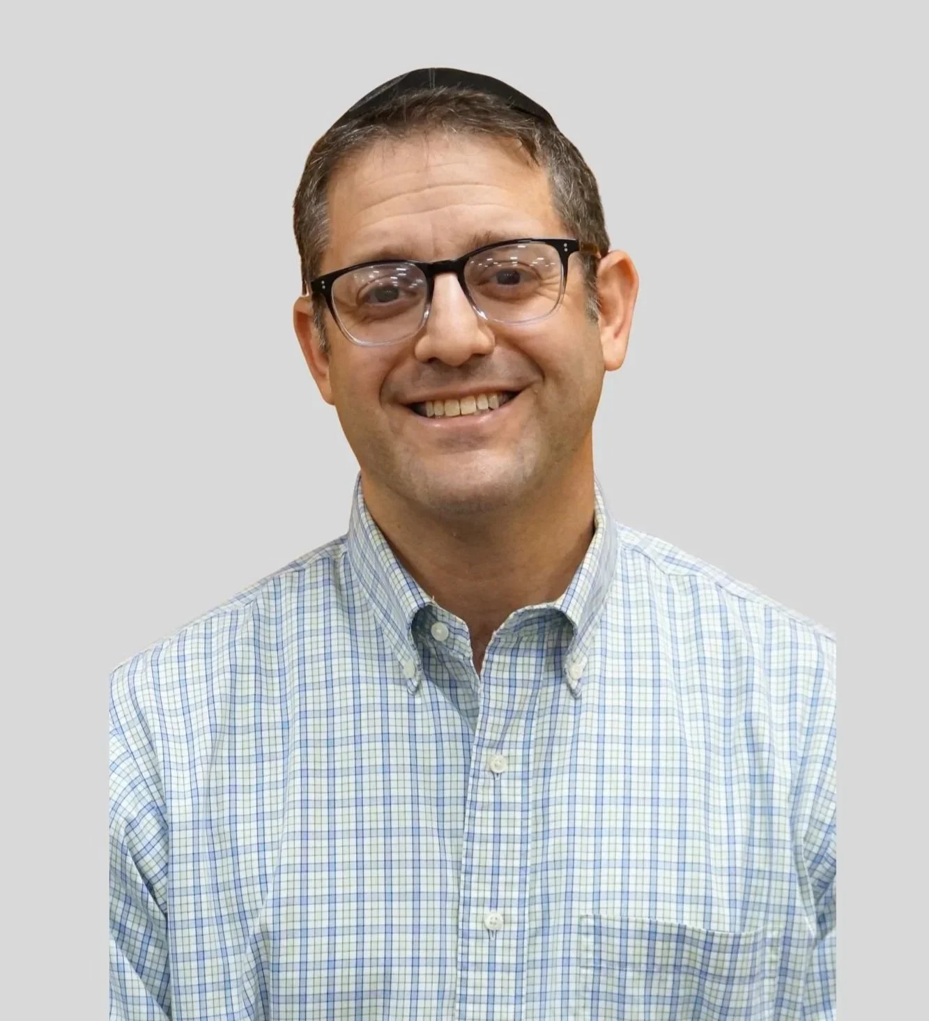 A smiling man wearing glasses and a checkered button-up shirt against a plain gray background.