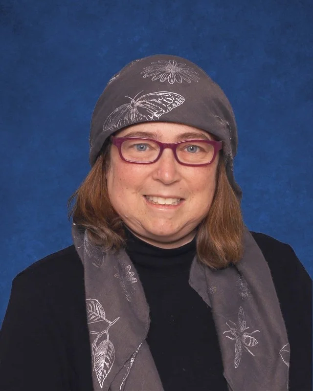 A woman with shoulder-length brown hair, purple glasses, and a dark gray headscarf with white floral and butterfly patterns, wearing a black top, smiles in front of a blue background.