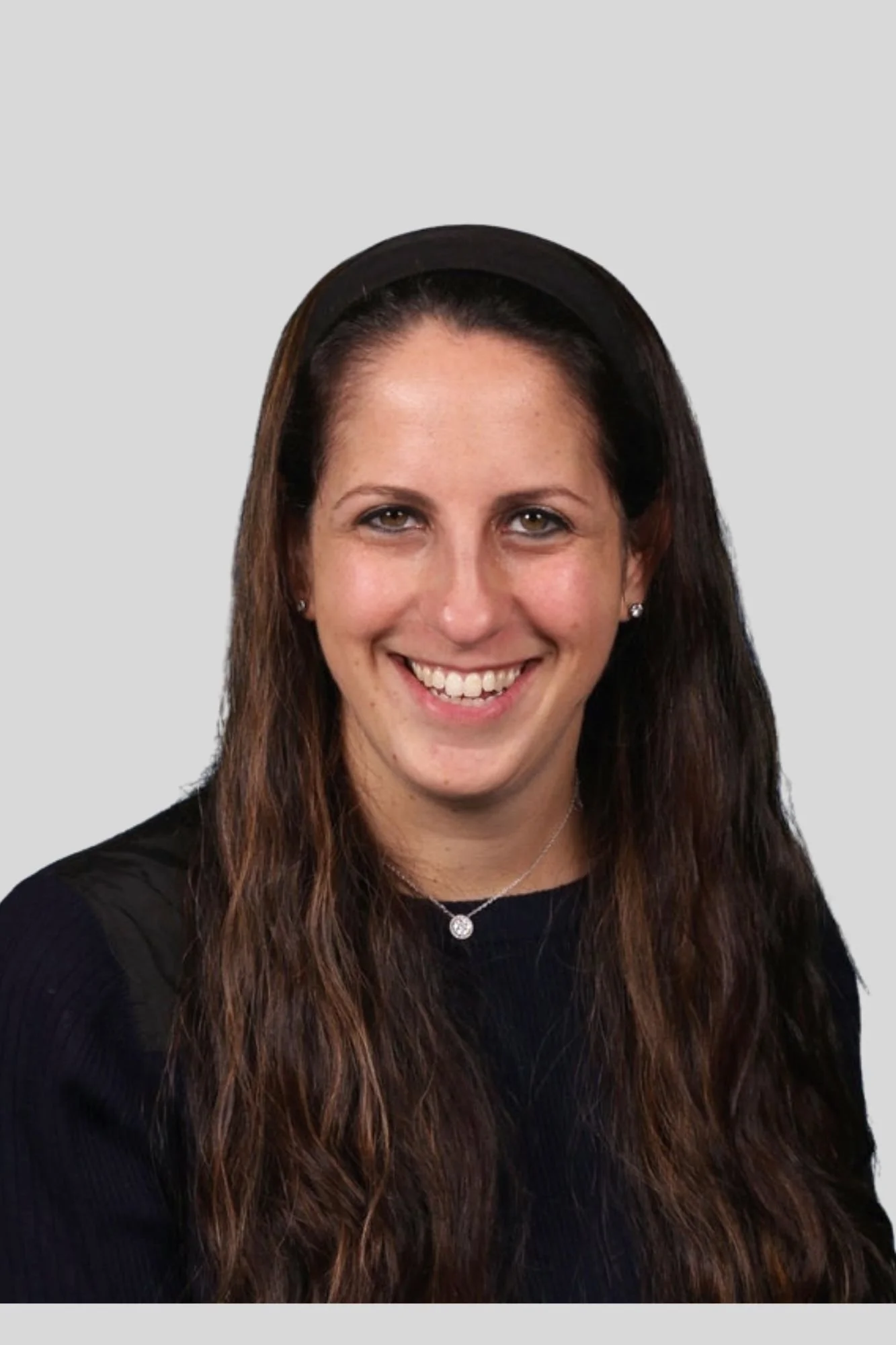 A woman with long brown hair wearing a black headband, black shirt, and jewelry, smiling against a light gray background.