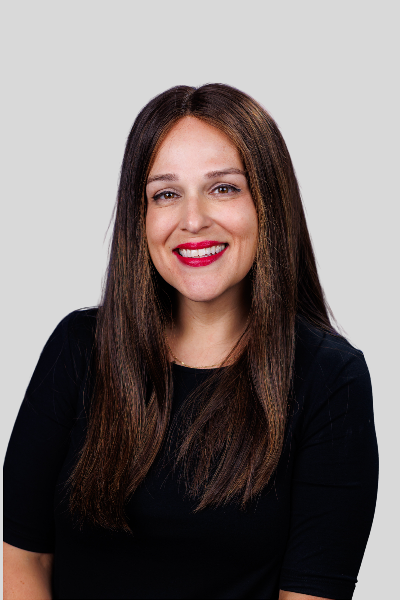 A woman with long brown hair and freckles smiling at the camera, wearing a black top and red lipstick against a plain light gray background.