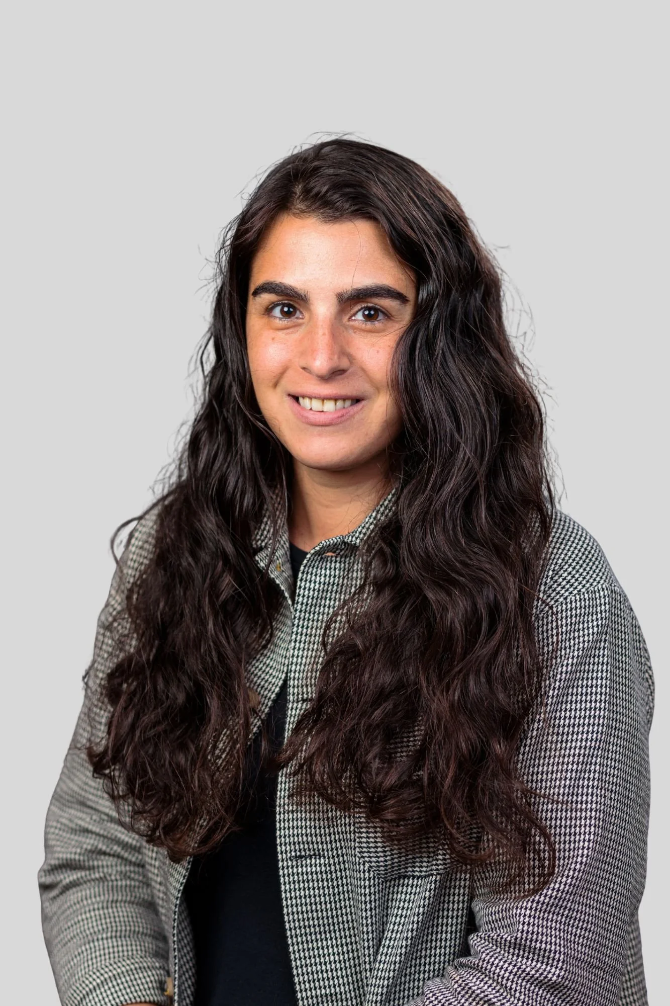A young woman with long, dark, curly hair and brown eyes smiling, wearing a black top and a checkered blazer, against a plain gray background.