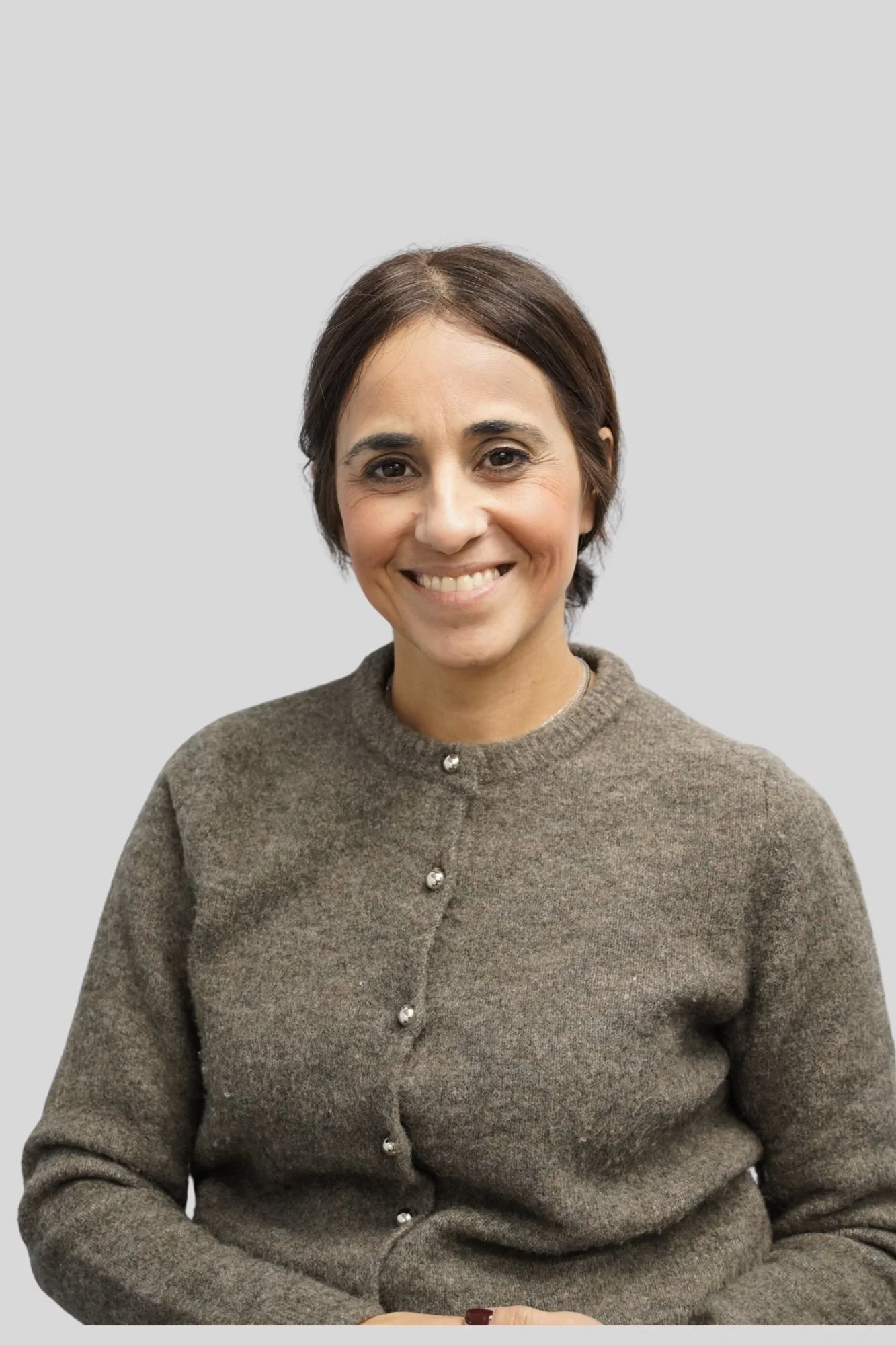 A woman with short brown hair, smiling, wearing a brown sweater with buttons, against a light gray background.