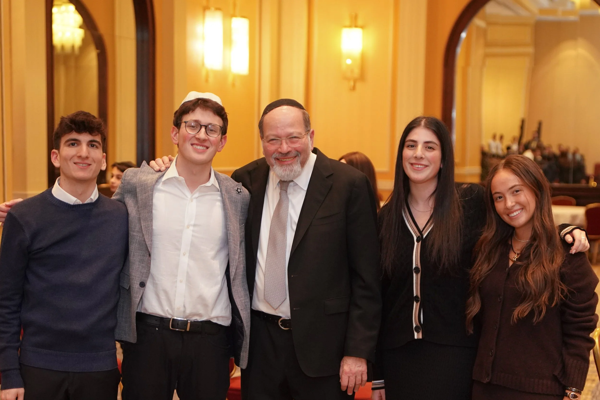 Group of five people smiling and posing for a photo in a well-lit room with yellow walls, including a man with glasses and a beard in the center, surrounded by two men and two women.
