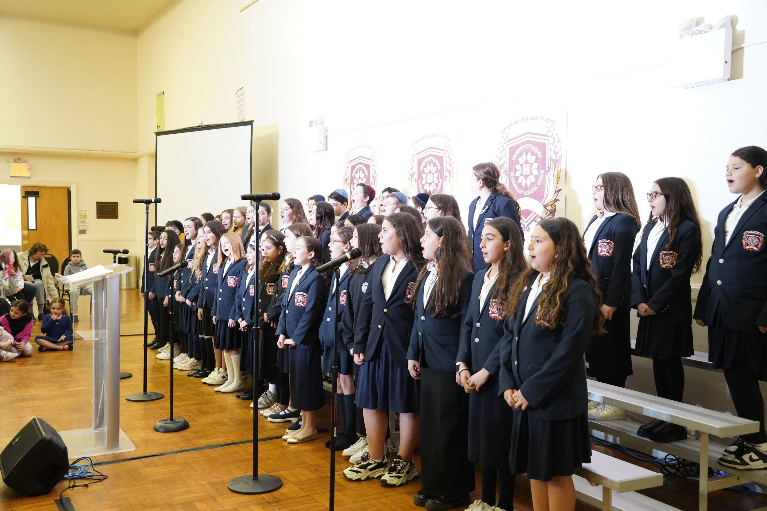 A group of children in uniform singing on stage during a school event in a gymnasium or auditorium. Many children are standing in rows, and there are microphones set up in front of them.