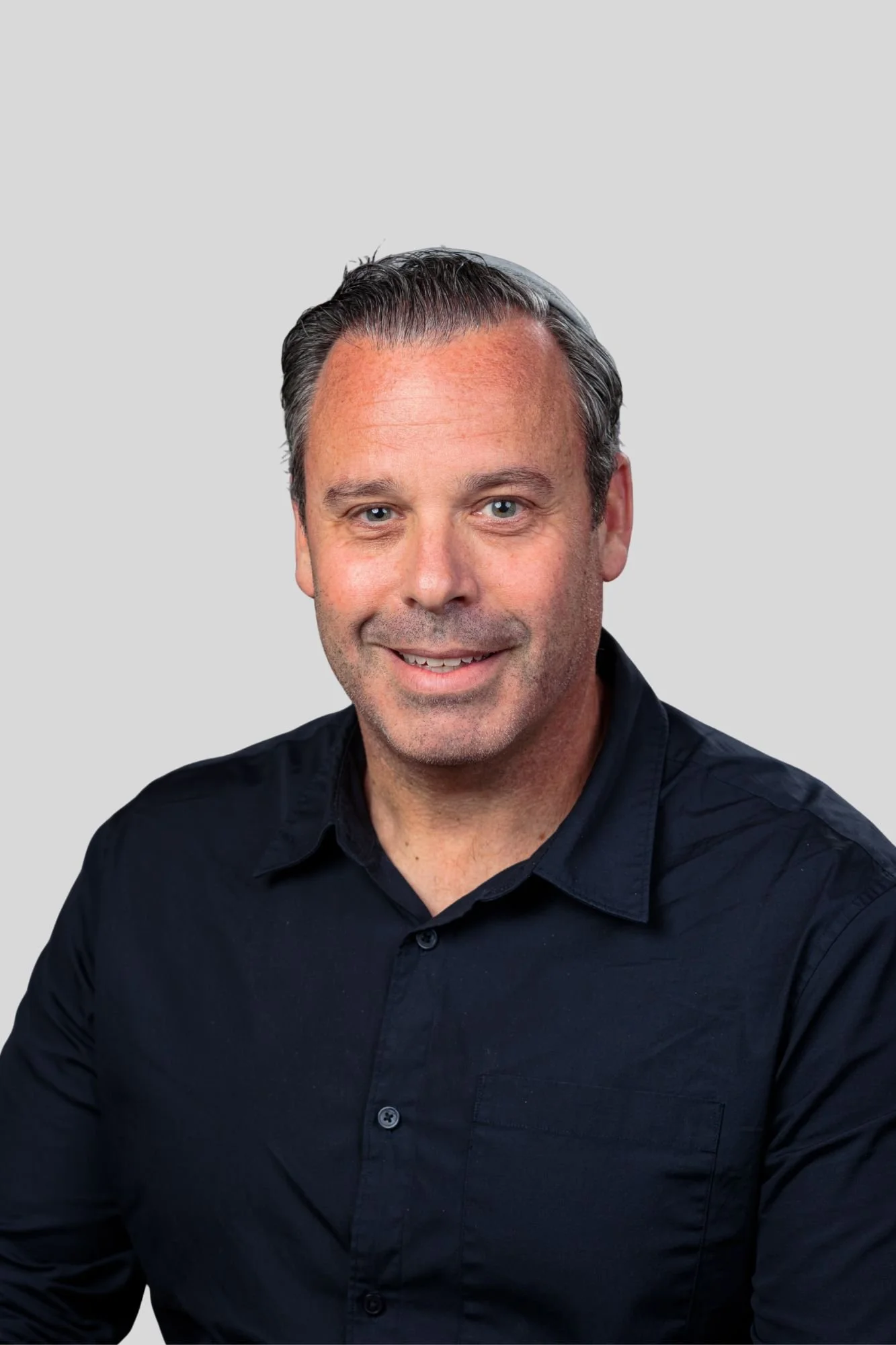 A headshot of a man smiling, wearing a black shirt, with short dark hair streaked with gray, and blue eyes, against a plain light gray background.