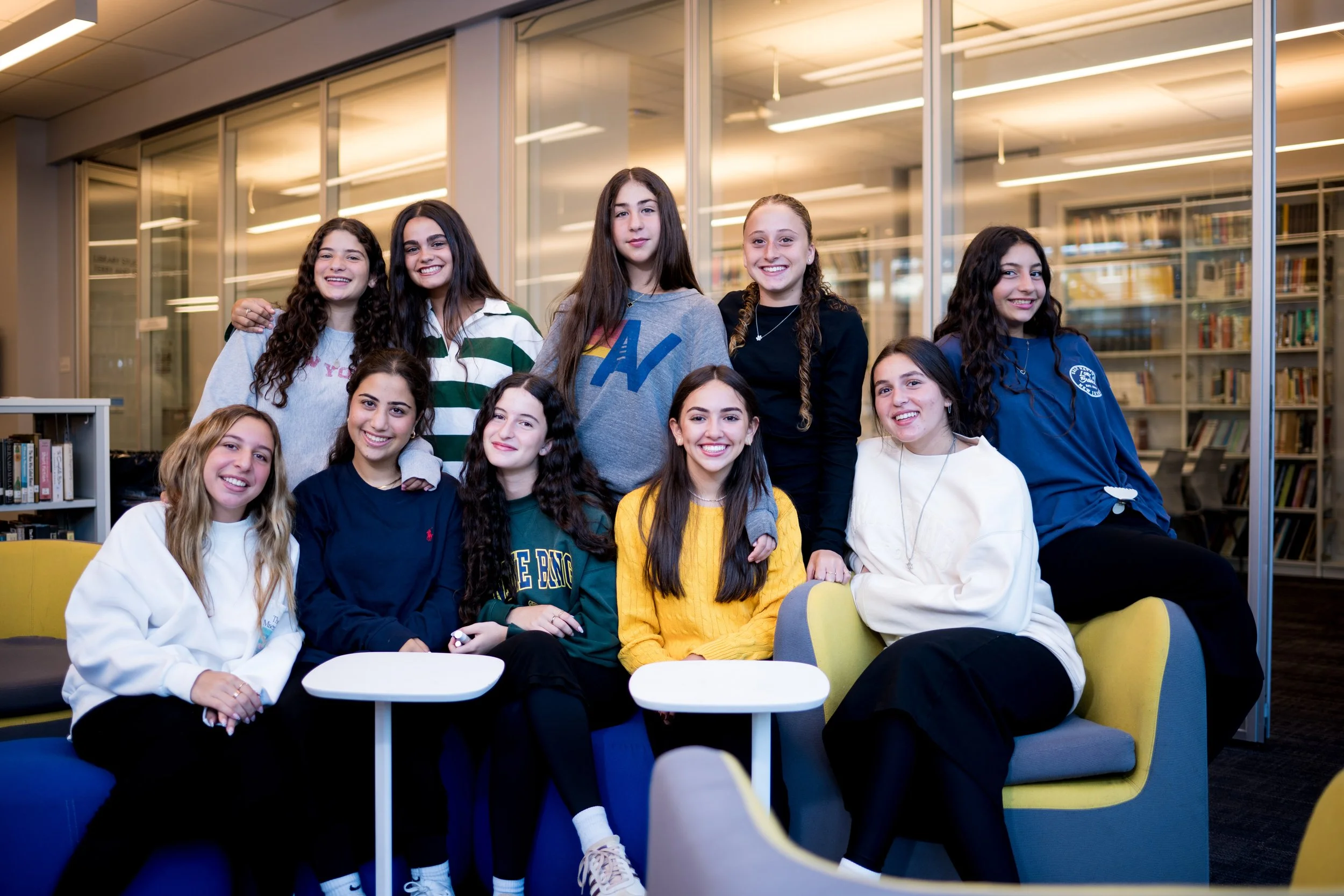 A group of ten teenage girls gathered in a library lounge area, some sitting on chairs and others standing behind, smiling at the camera.
