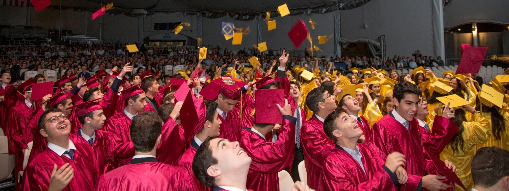 Graduate students dressed in red and yellow caps and gowns celebrating during graduation ceremony indoors, tossing caps into the air.