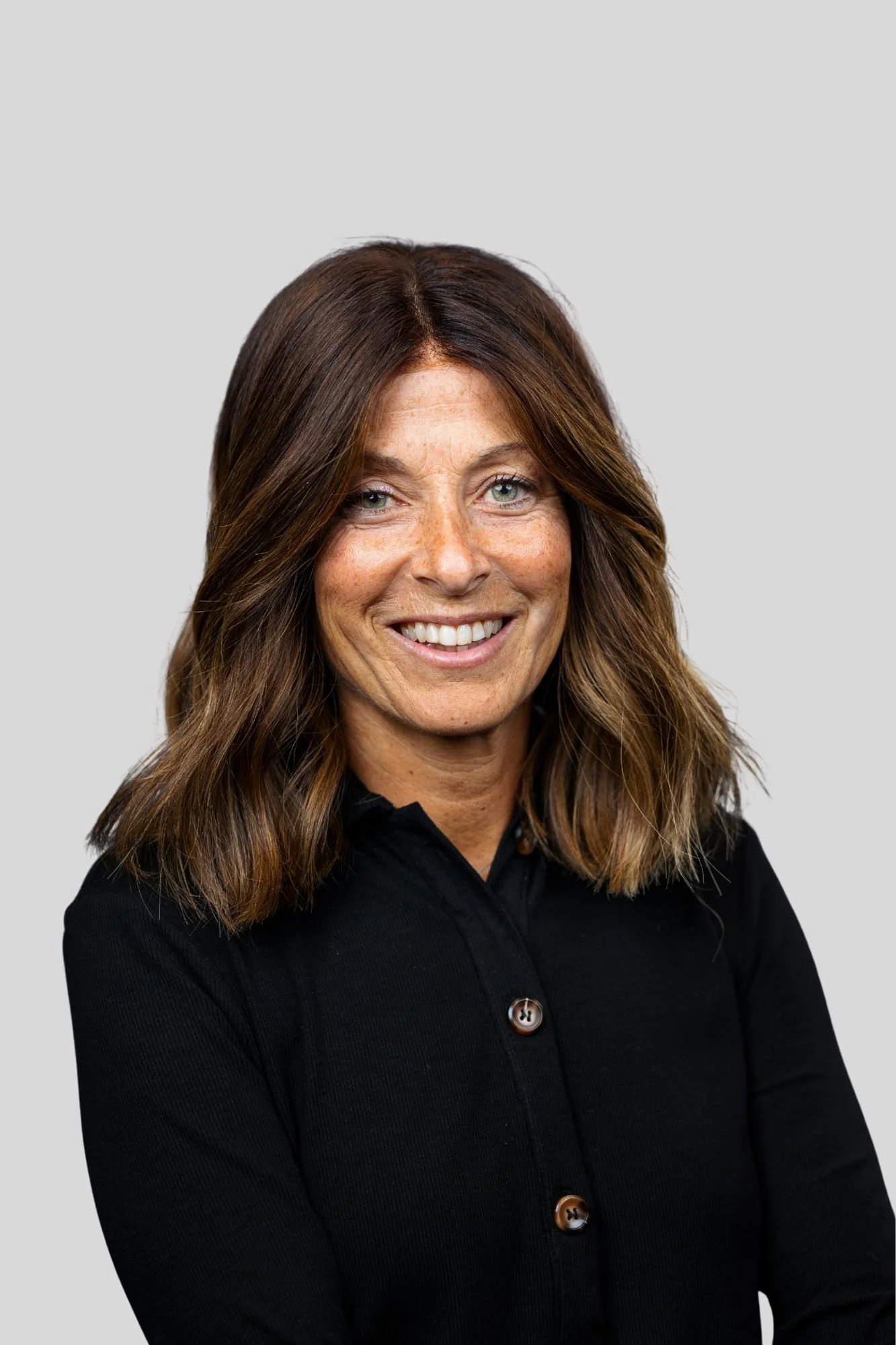 Close-up of a woman with long brown hair, light skin, and gray eyes, smiling, wearing a black button-up shirt, against a light gray background.