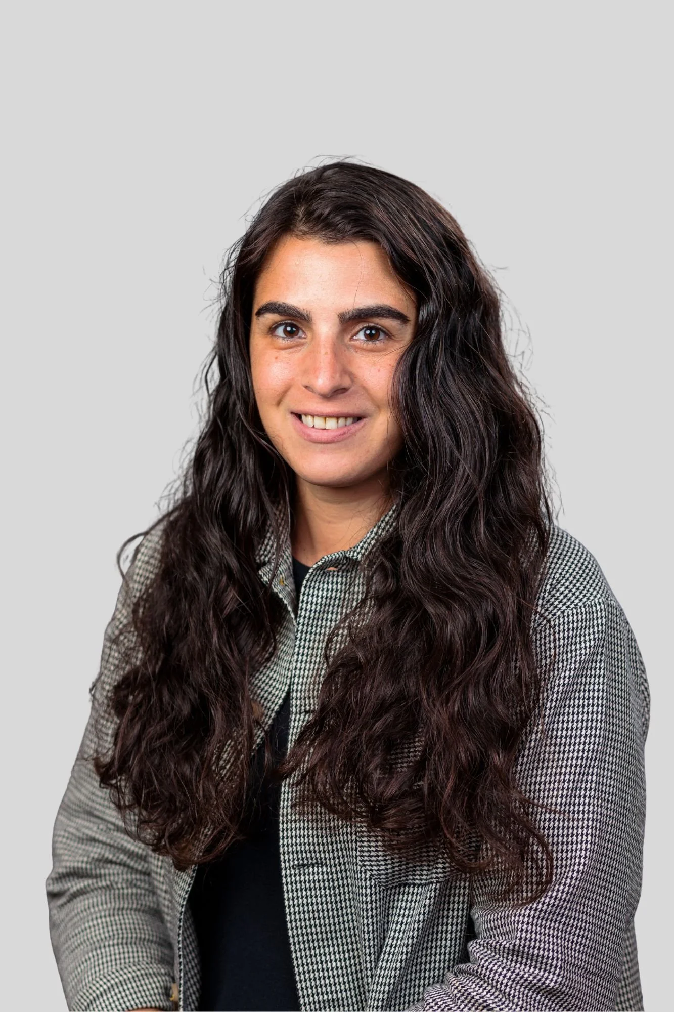 Portrait of a young woman with long, wavy dark hair, wearing a checkered blazer over a black top, smiling against a plain gray background.