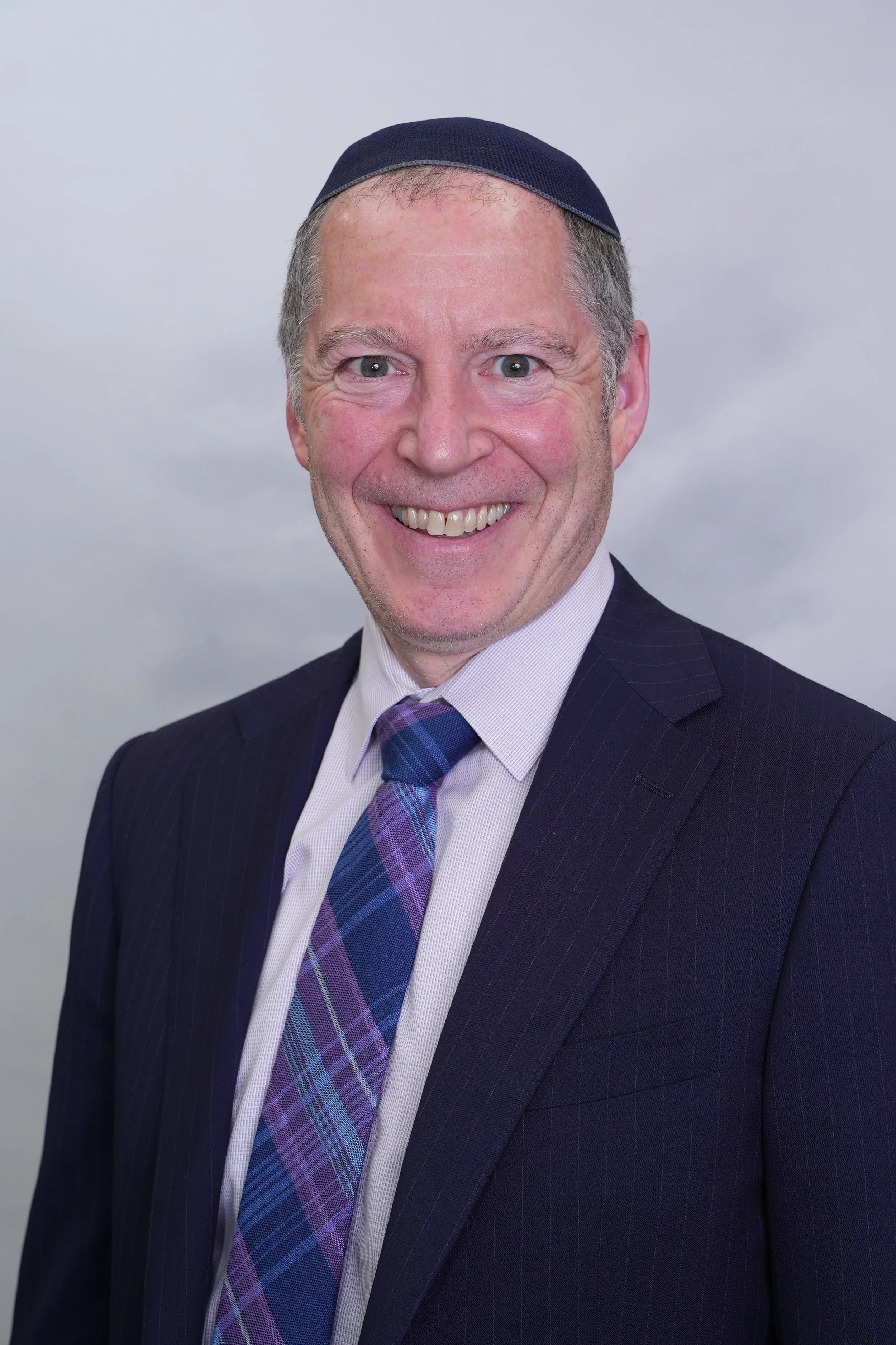 A smiling middle-aged man in a dark suit, white shirt, and plaid tie wearing a blue kippah, standing against a gray background.