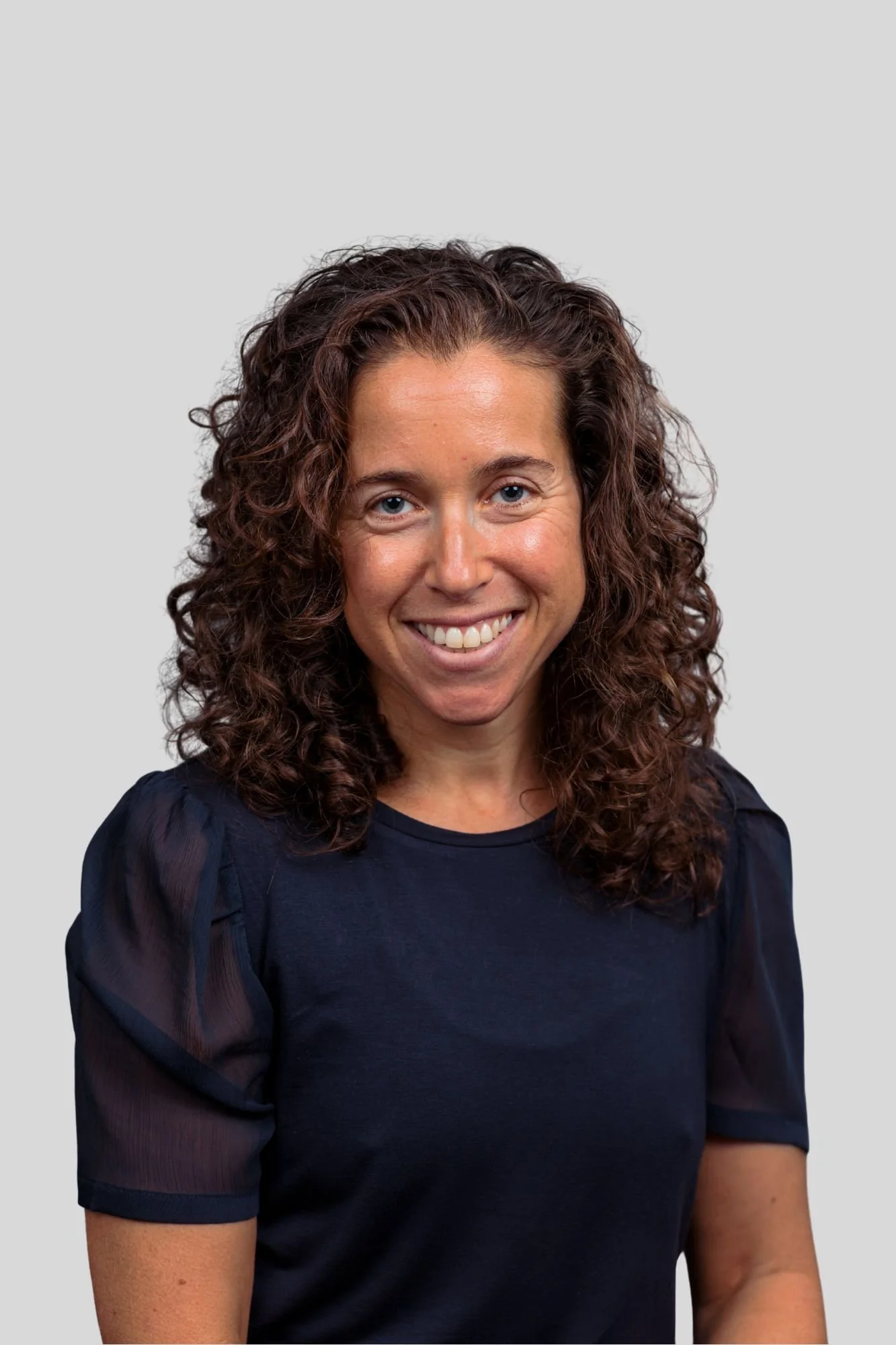 A woman with curly brown hair and blue eyes smiling, wearing a black top with sheer sleeves, standing against a plain gray background.