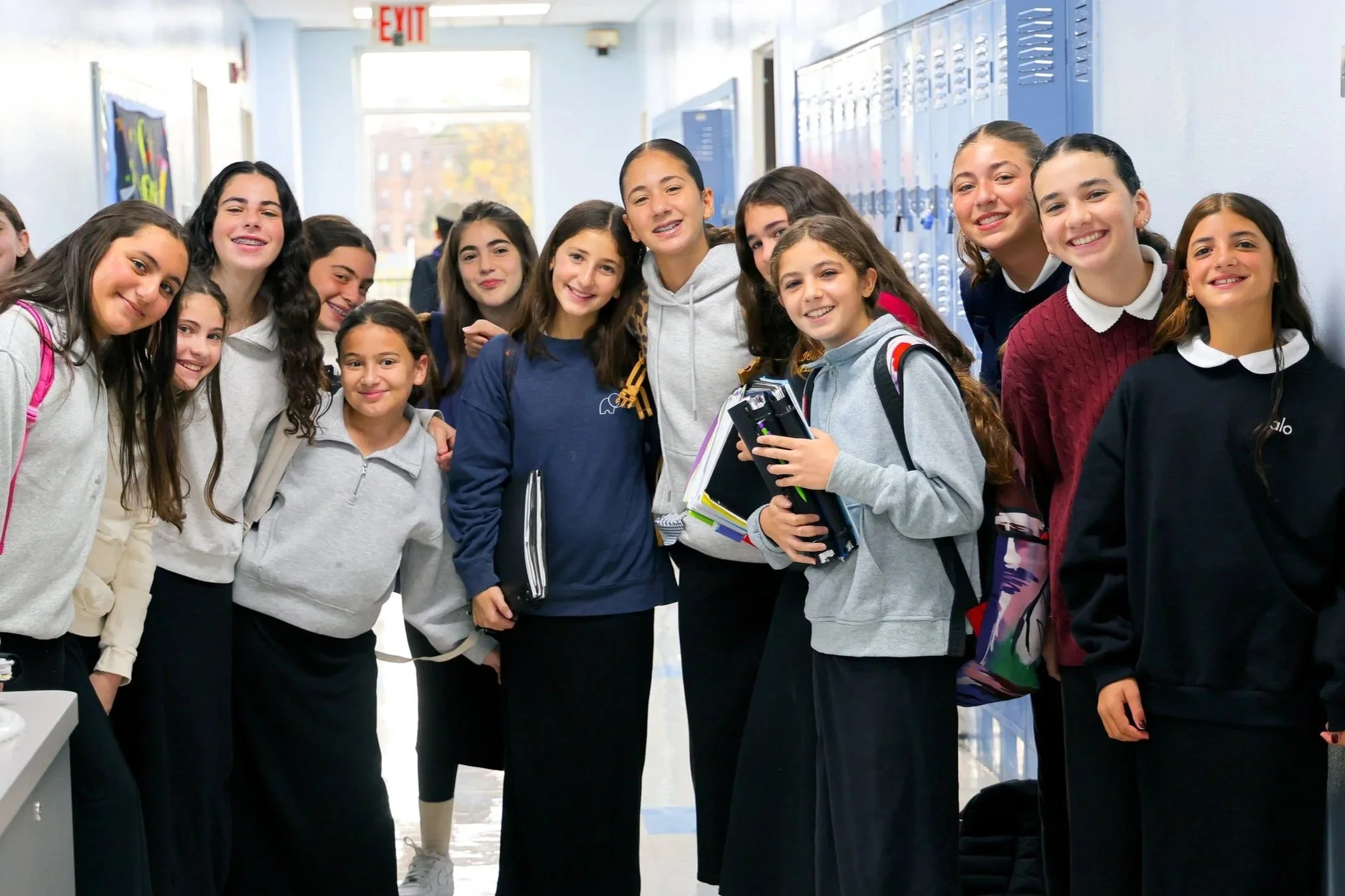 A group of middle school girls standing in a school hallway, smiling for a photo. They are wearing casual clothes and carrying backpacks and notebooks, with lockers and windows in the background.