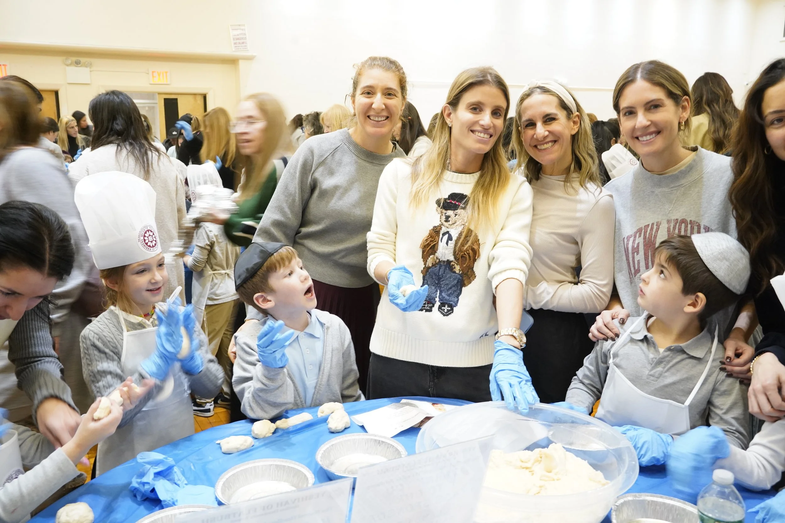 Group of women and children participating in a baking or cooking activity indoors, with baking trays and dough on a table covered with a blue tablecloth.