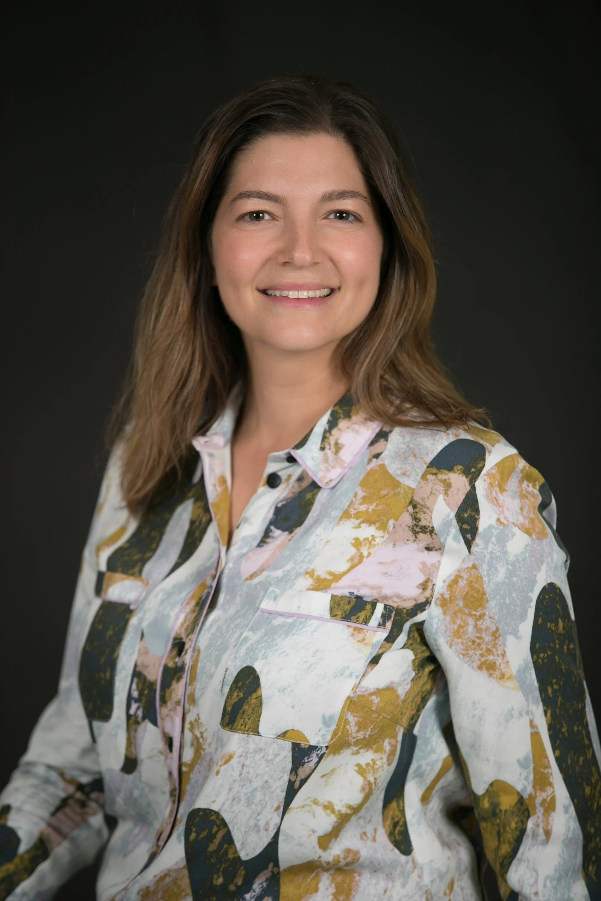 A woman with shoulder-length brown hair smiling at the camera, wearing a floral-patterned button-up shirt, against a dark background.