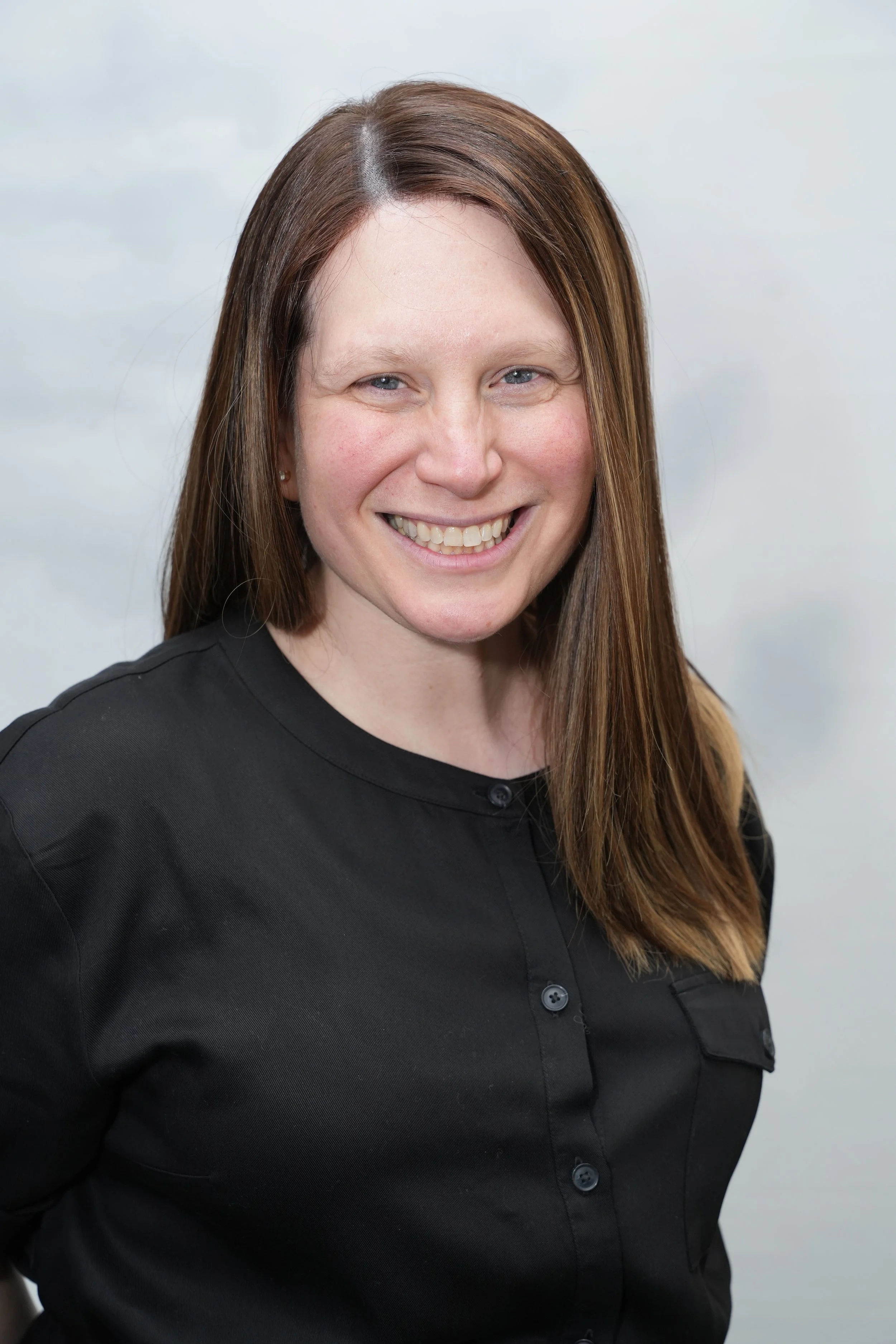 Close-up of a woman with long brown hair wearing a black button-up shirt, smiling against a neutral background.
