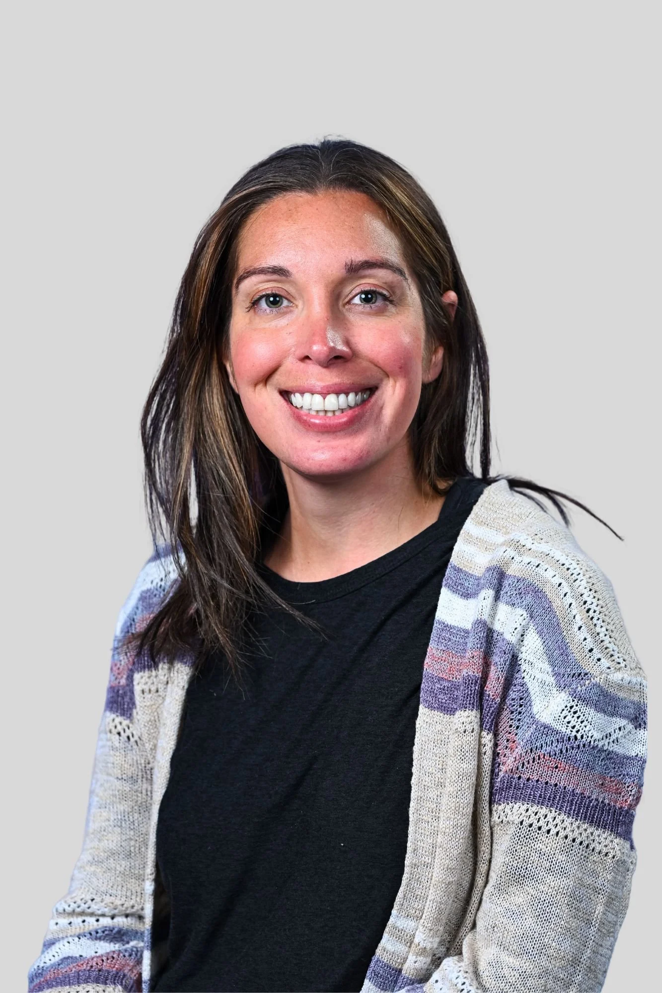 A smiling woman with shoulder-length brown hair, wearing a black top and a multicolored knit cardigan, standing against a plain light gray background.