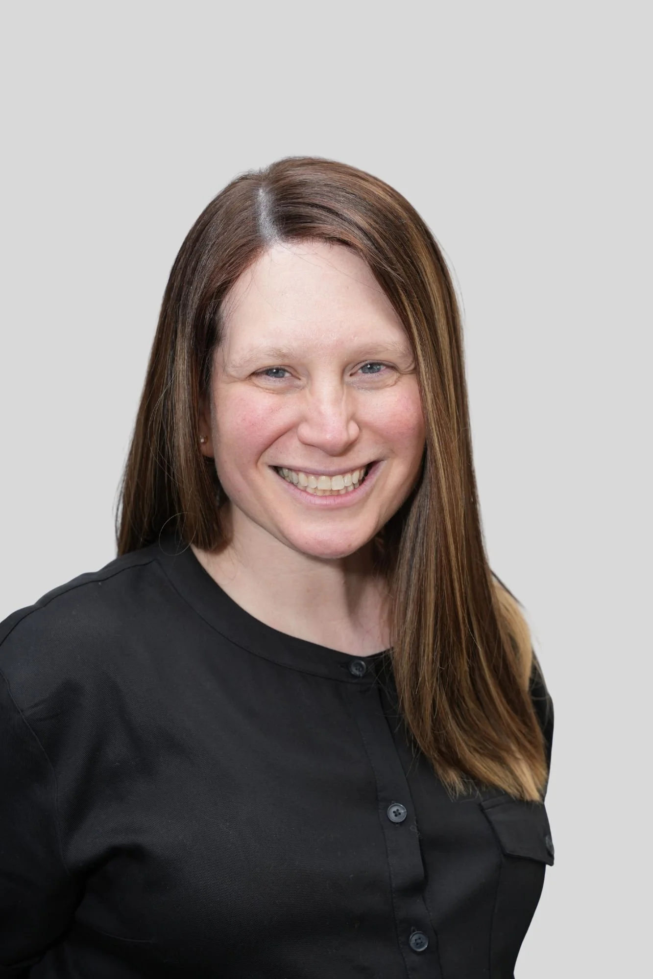 A woman with straight, long, light brown hair smiling, wearing a black collared shirt, against a plain light gray background.
