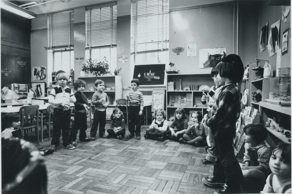Group of young children in a classroom, some standing and some sitting on the floor, with artwork and shelves in the background.