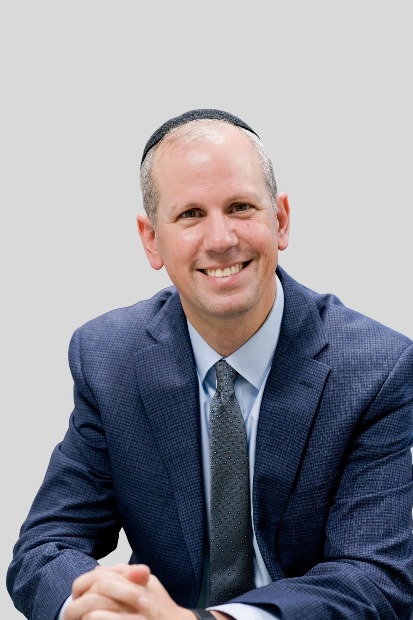 A smiling man wearing a dark kippah, navy blue checkered suit, light blue shirt, and grey polka dot tie, sitting with hands clasped.