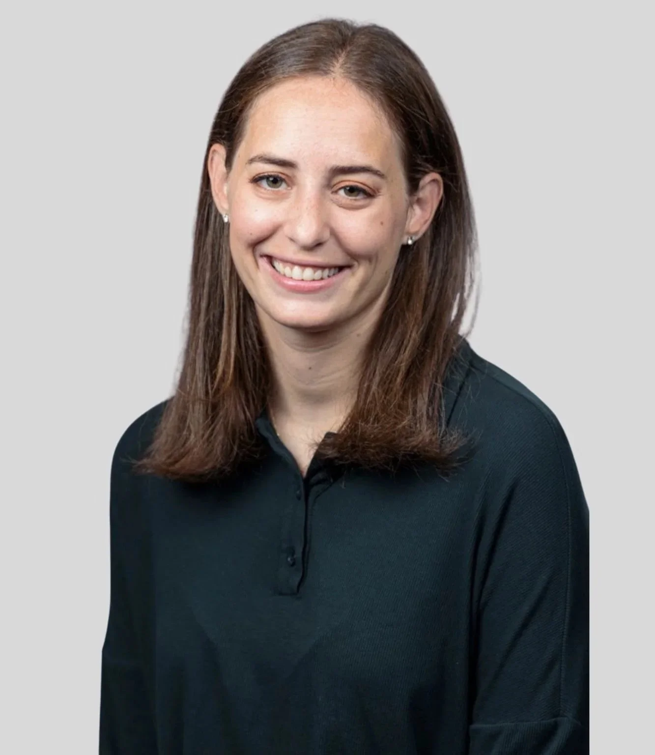 A woman with shoulder-length brown hair, smiling, wearing a black collared shirt, against a light gray background.
