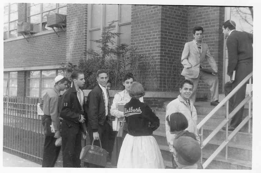 A group of young people standing outside a school building, waiting to enter, with some on the stairs and others on the sidewalk.