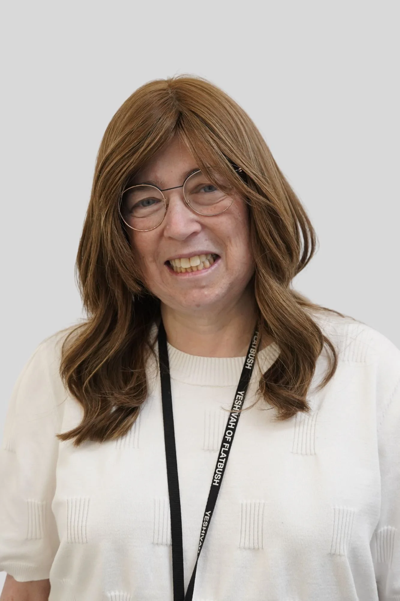A woman with long, wavy, light brown hair, wearing glasses and a white sweater, smiling in front of a plain light gray background. She has a black lanyard with white text around her neck.