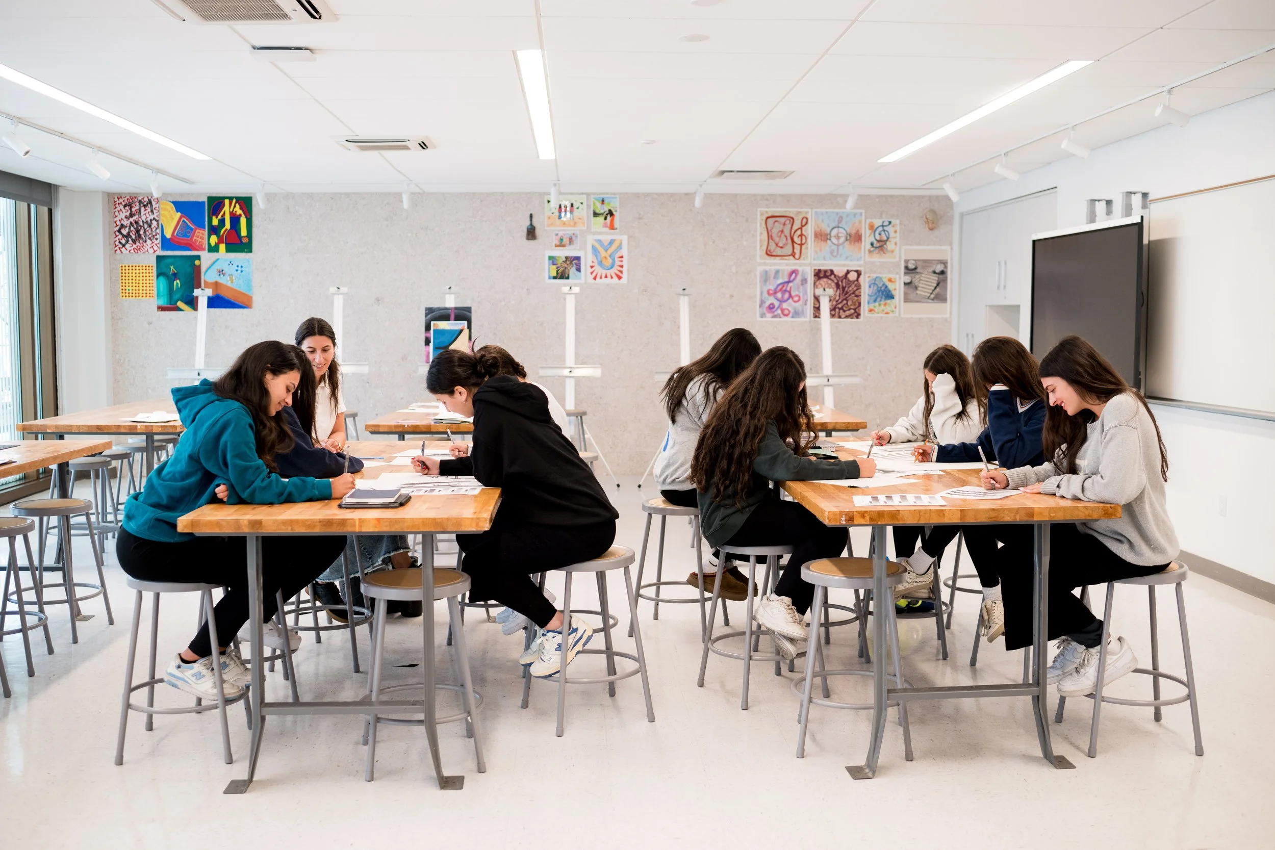 A classroom with students sitting at tables, writing or working on papers, and colorful artwork hanging on the walls.