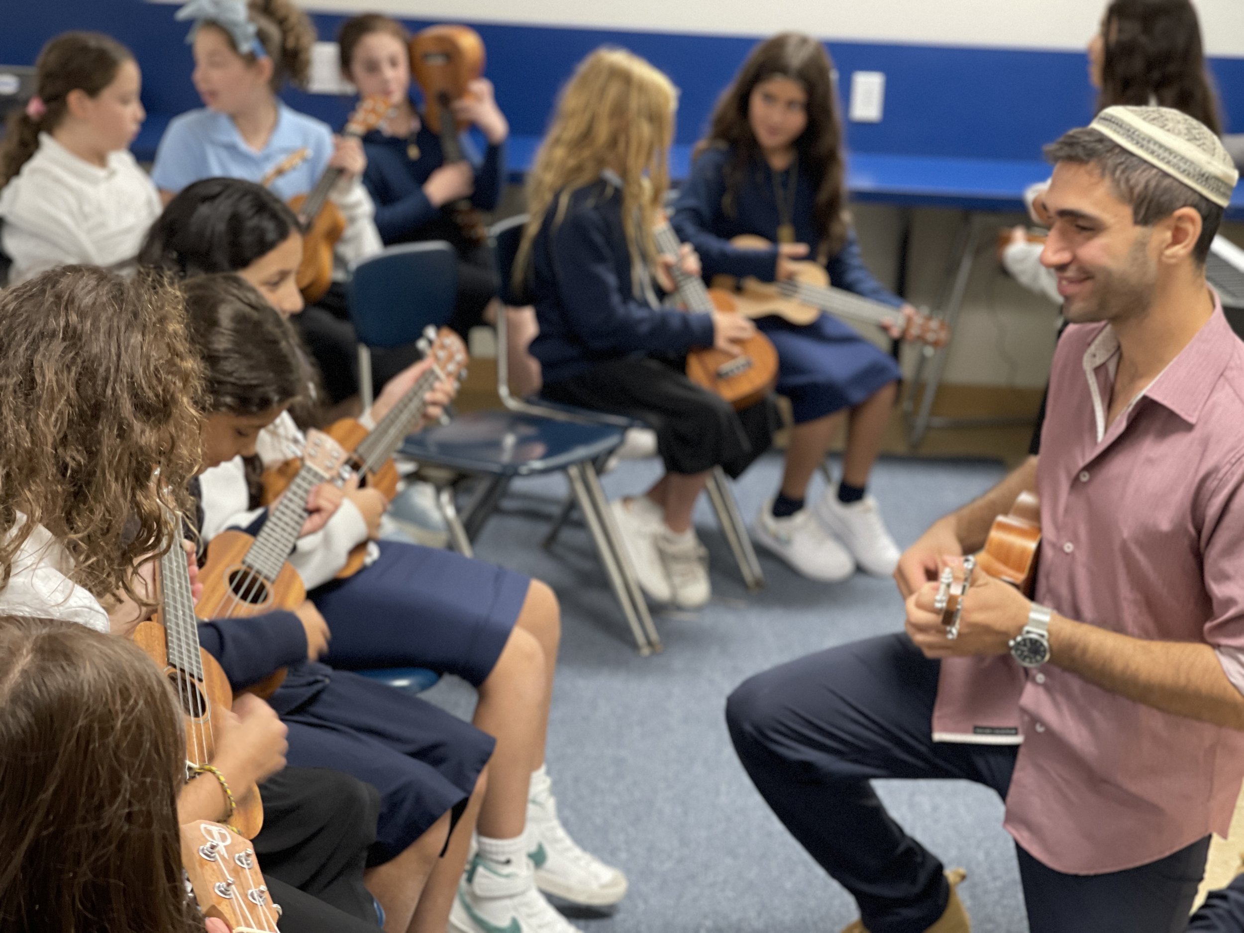 Group of children sitting on chairs, playing small ukuleles in a classroom, while an adult male instructor, wearing a yarmulke and a watch, is kneeling and teaching them.