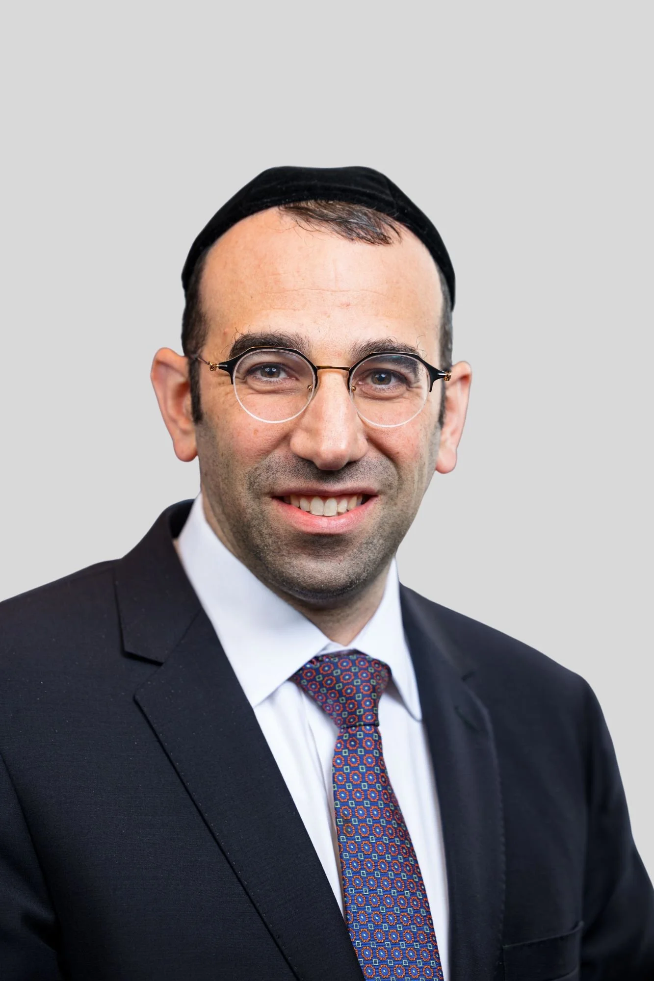 Portrait of a man wearing glasses, a yarmulke, a dark tuxedo, a white shirt, and a patterned tie, smiling against a plain gray background.