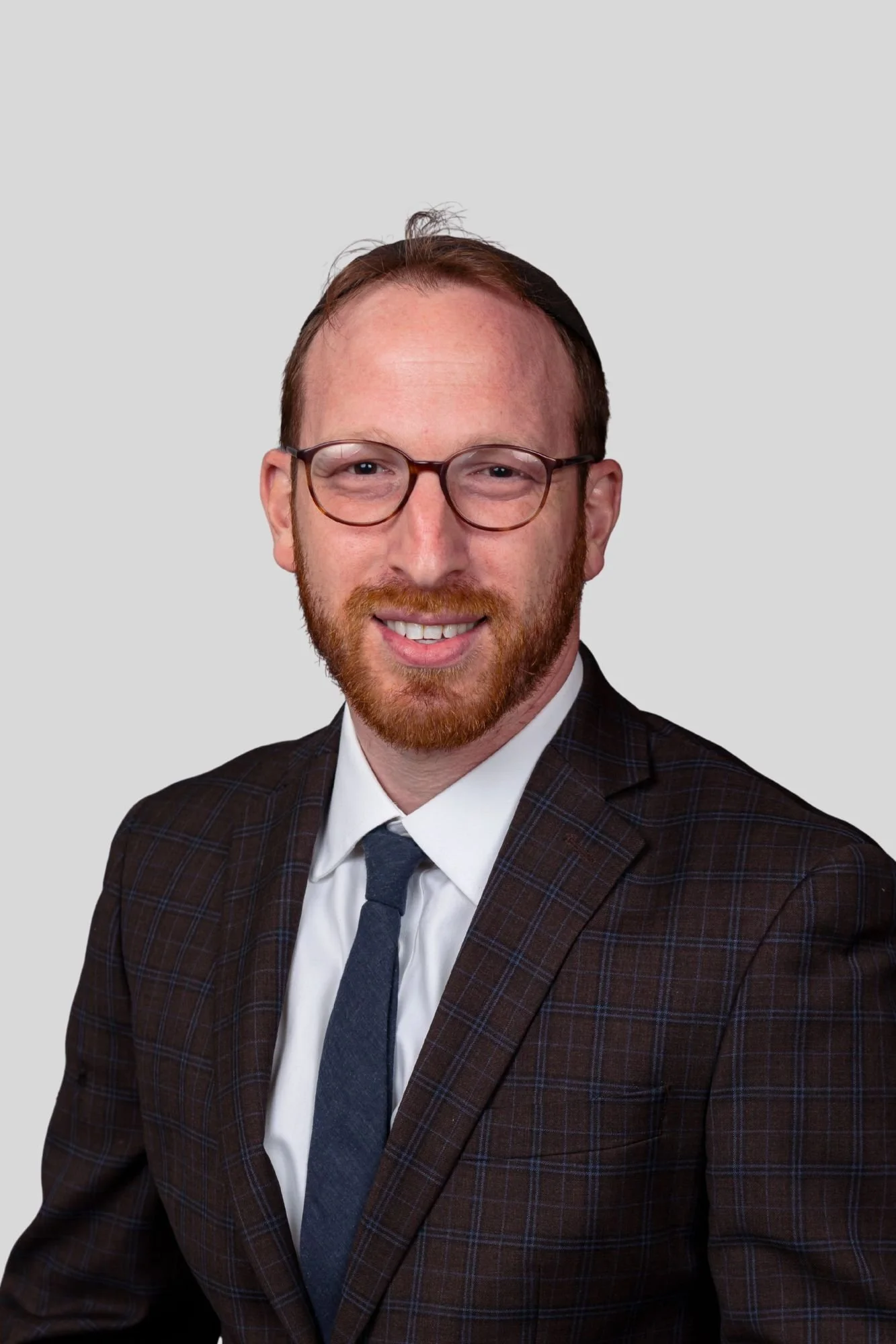 A man wearing glasses, a white shirt, a blue tie, and a dark plaid suit against a light gray background.