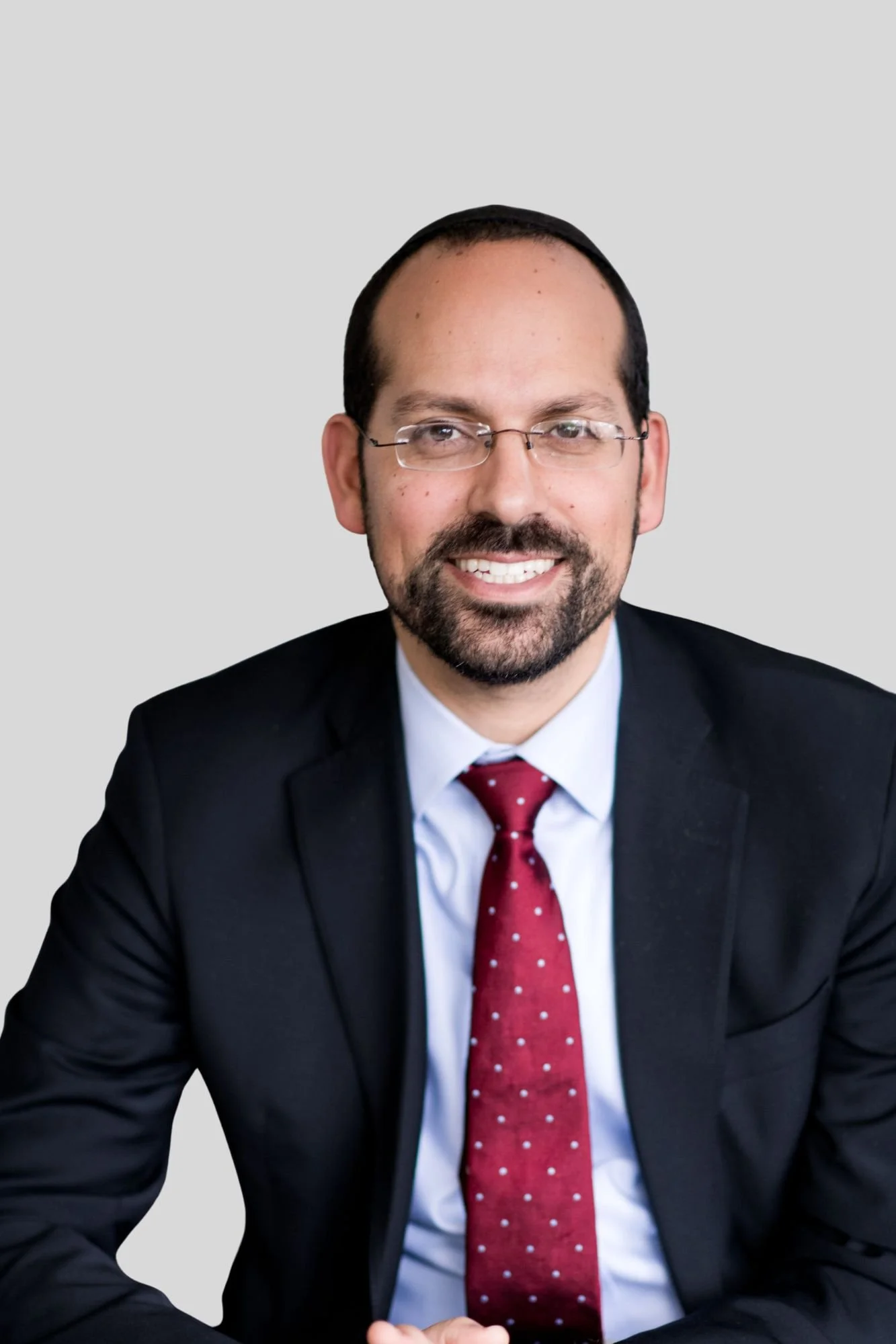 A man with glasses, dark hair, and a beard smiling, wearing a black suit, white shirt, and red tie with white polka dots, seated against a plain light gray background.