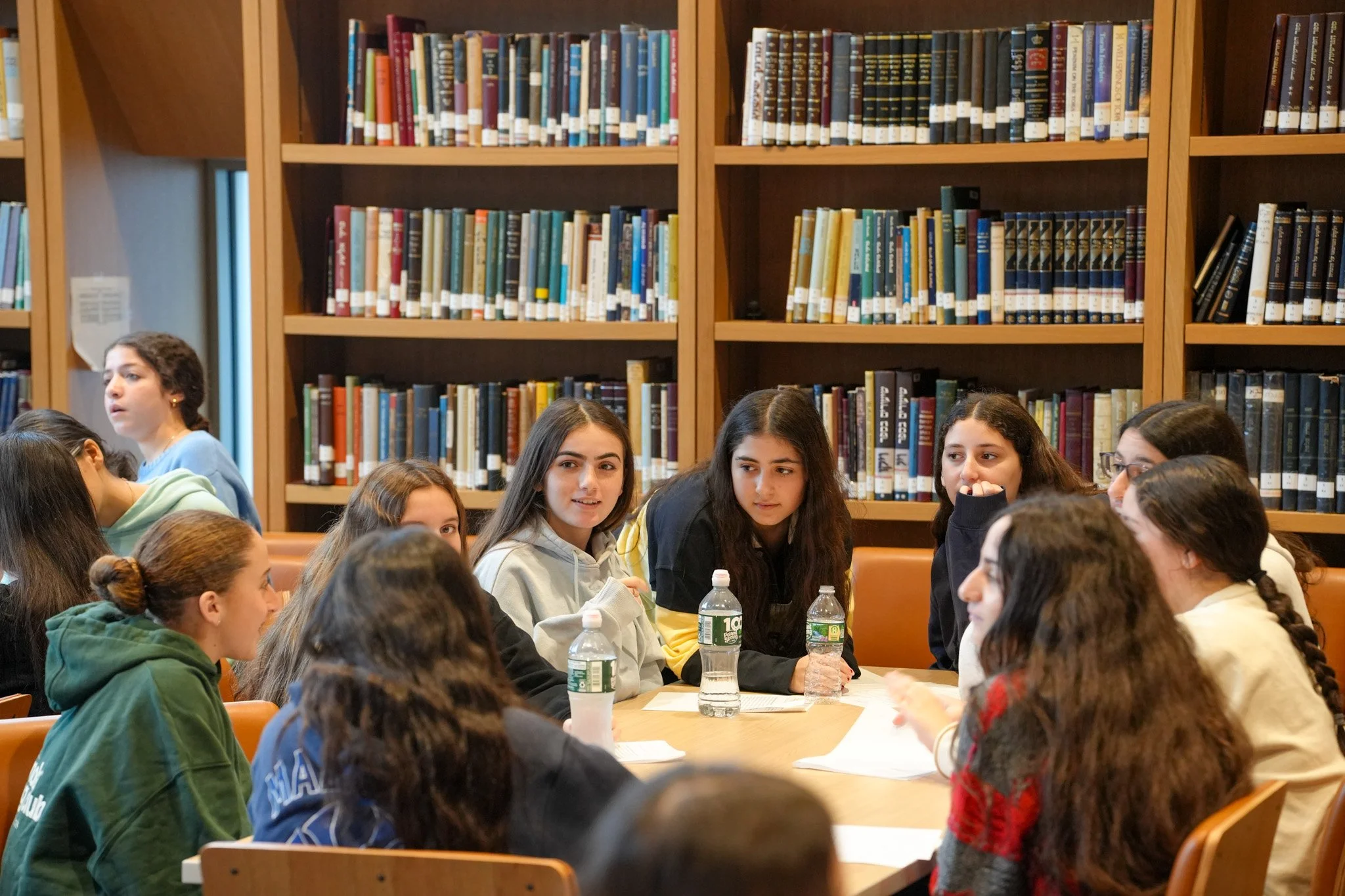 Group of students and teacher in a library classroom having a discussion, with shelves of books behind them.