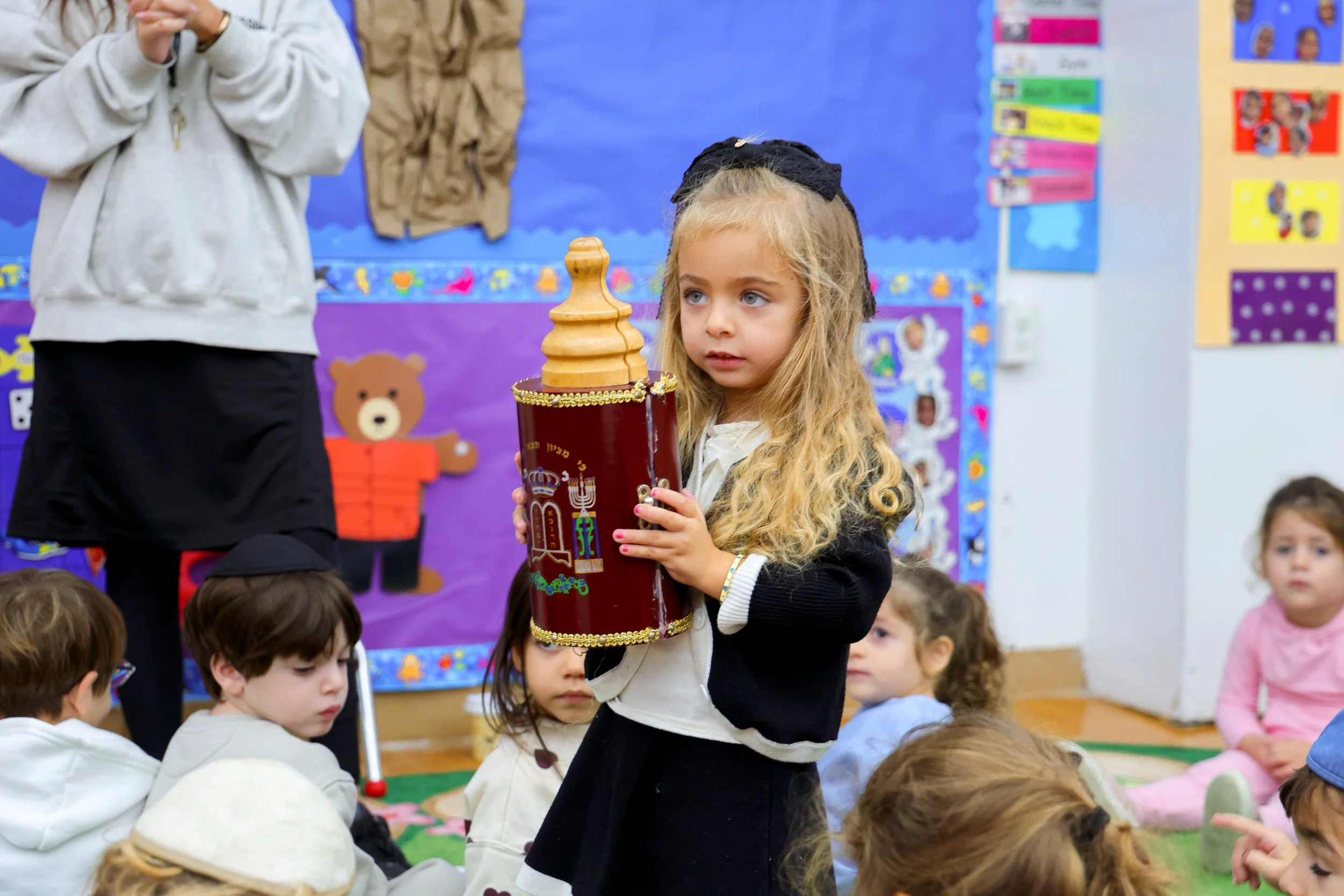 A young girl with long blonde hair holding a large binoculars with the Israeli flag design while standing in a classroom filled with children, colorful posters, and decorations.