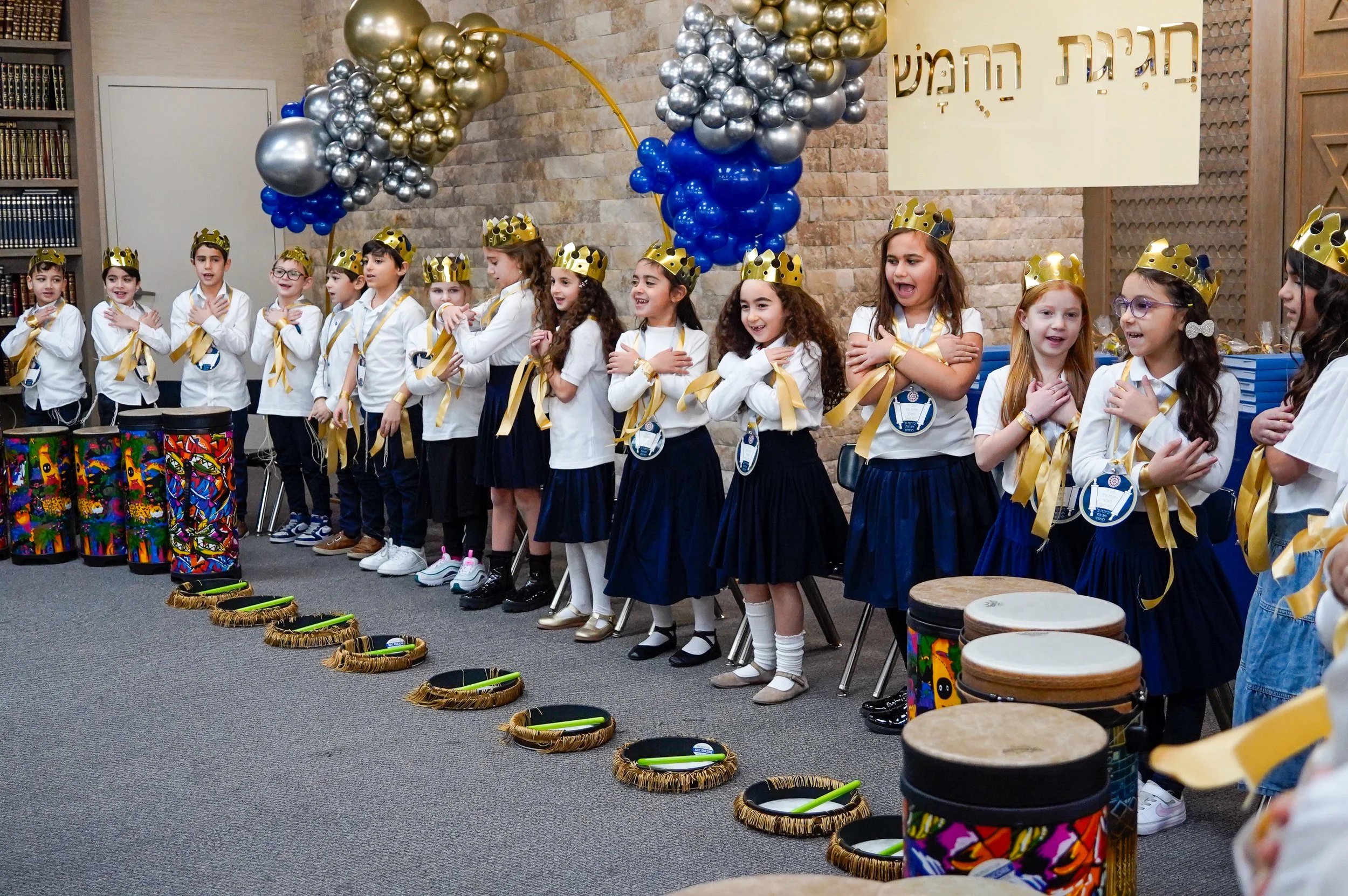 Children wearing gold crowns stand in a line indoors with a brick wall and Hebrew writing behind them. They are participating in a ceremony, with some placing their right hands on their chests. There are drums in front of them and a colorful balloon arrangement overhead.