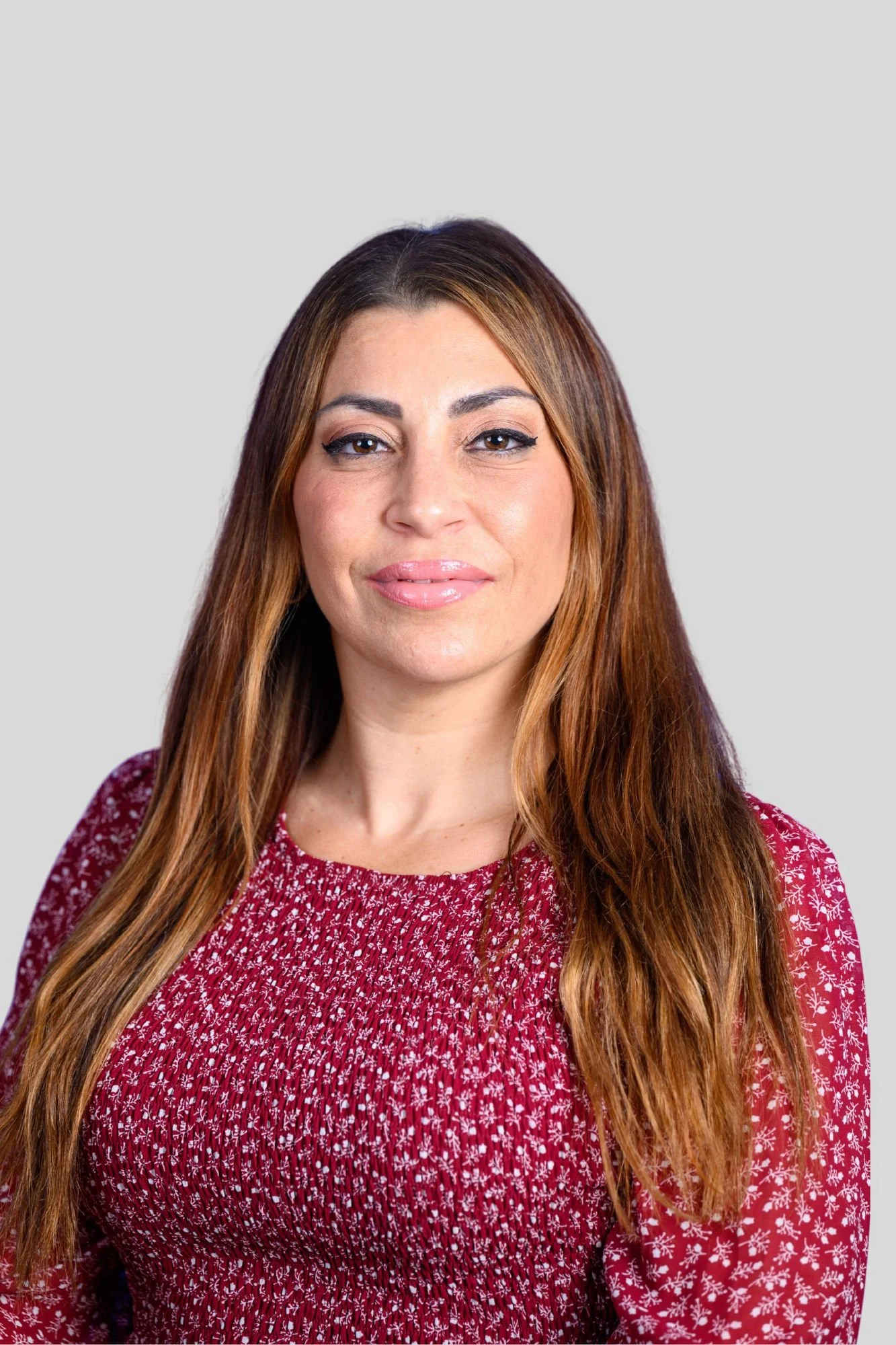 A woman with long brown hair, wearing a red patterned dress and standing against a plain grey background.