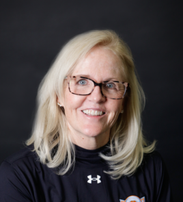 A woman with blonde hair wearing glasses and a black Under Armour shirt, smiling against a dark background.