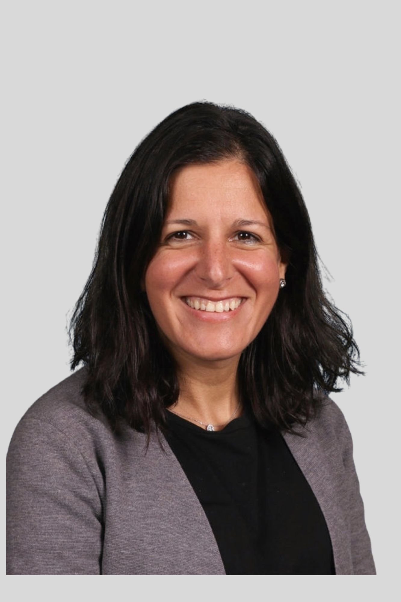 A woman with dark hair smiling, wearing a gray blazer over a black top, in front of a light gray background.