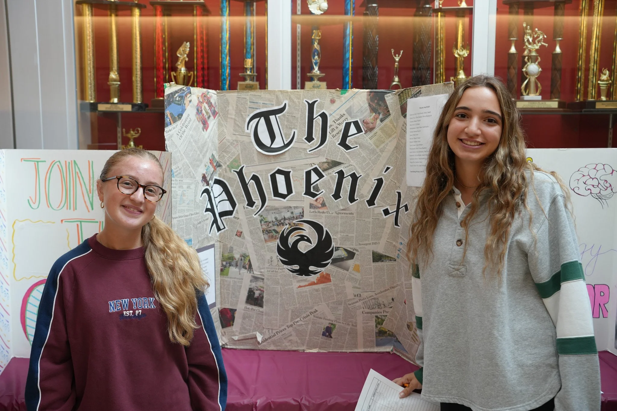 Two young girls standing in front of a display board with a backdrop of trophies. One girl is wearing glasses and a maroon sweatshirt, and the other has long wavy hair and is wearing a gray hoodie. They are smiling at the camera.