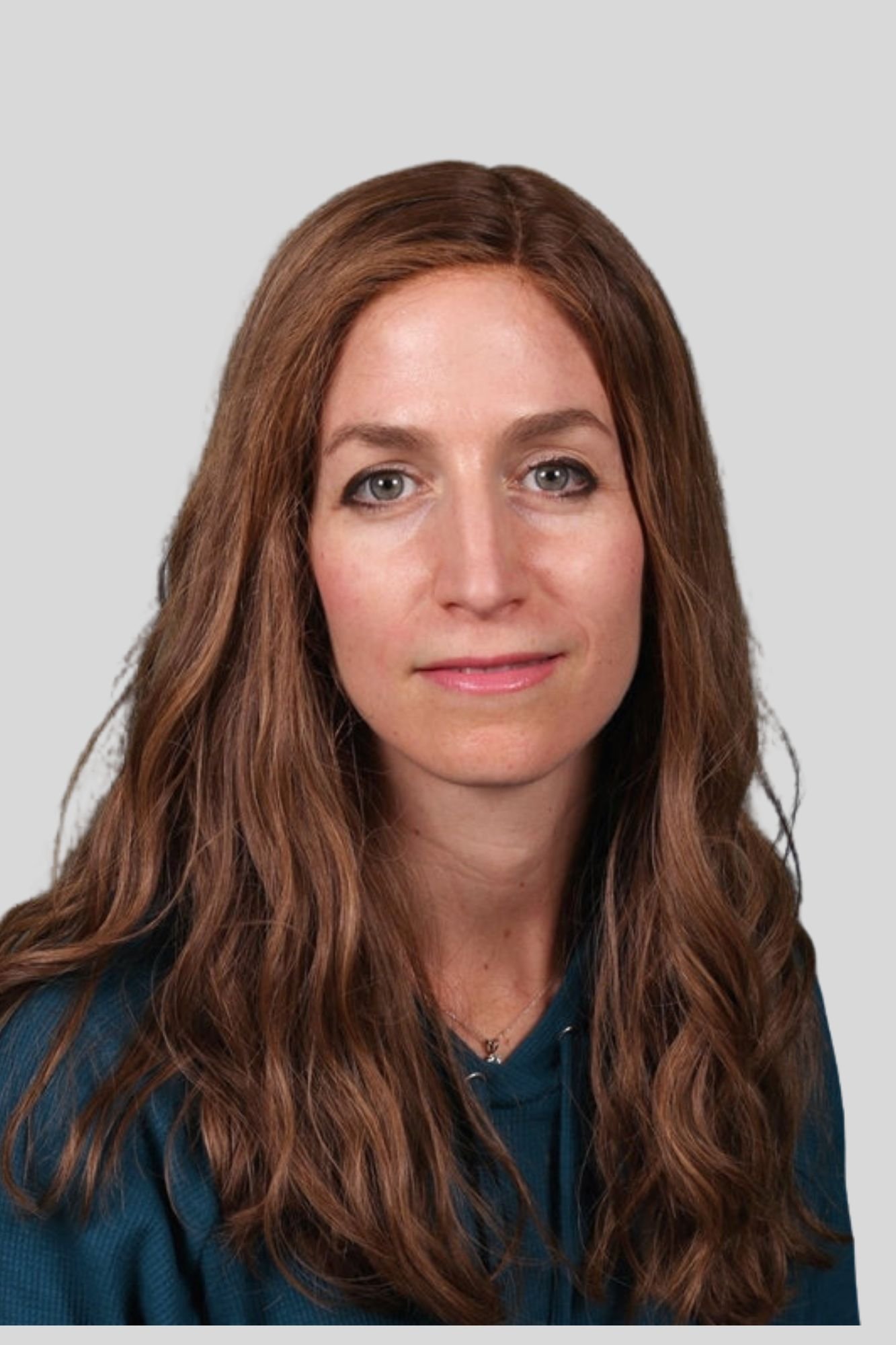 A woman with long wavy auburn hair, blue eyes, and a subtle smile, wearing a dark blue top and a gold necklace, against a plain light gray background.